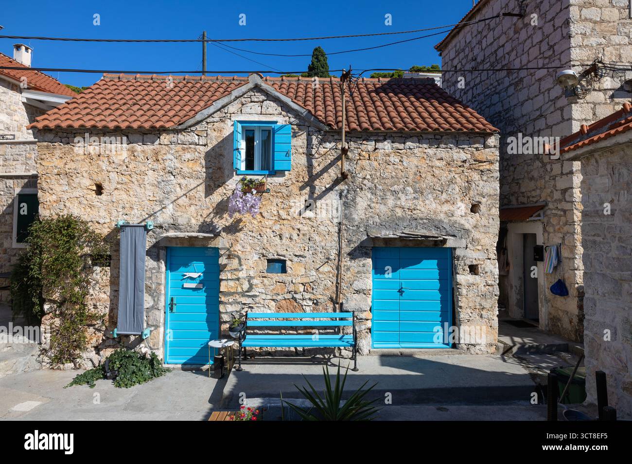 Una casa in pietra intemprata con porte e persiane blu vibranti si trova su una strada mediterranea illuminata dal sole. Ivy si arrampica sul muro, una panchina blu aggiunge fascino, e. Foto Stock