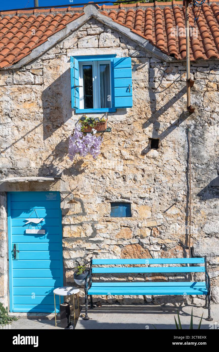 Una casa in pietra intemprata con porte e persiane blu vibranti si trova su una strada mediterranea illuminata dal sole. Ivy si arrampica sul muro, una panchina blu aggiunge fascino, e. Foto Stock