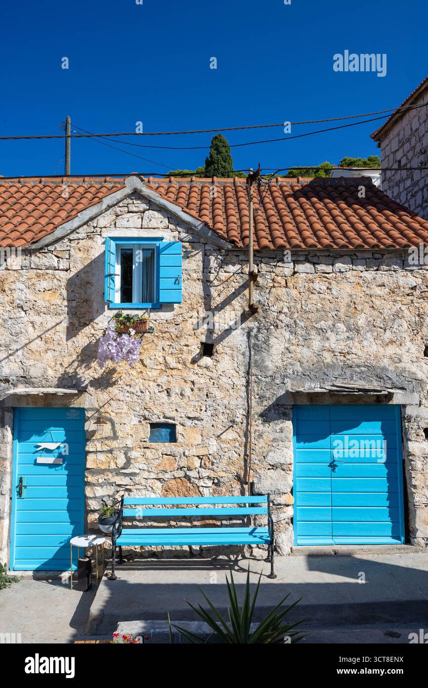 Una casa in pietra intemprata con porte e persiane blu vibranti si trova su una strada mediterranea illuminata dal sole. Ivy si arrampica sul muro, una panchina blu aggiunge fascino, e. Foto Stock