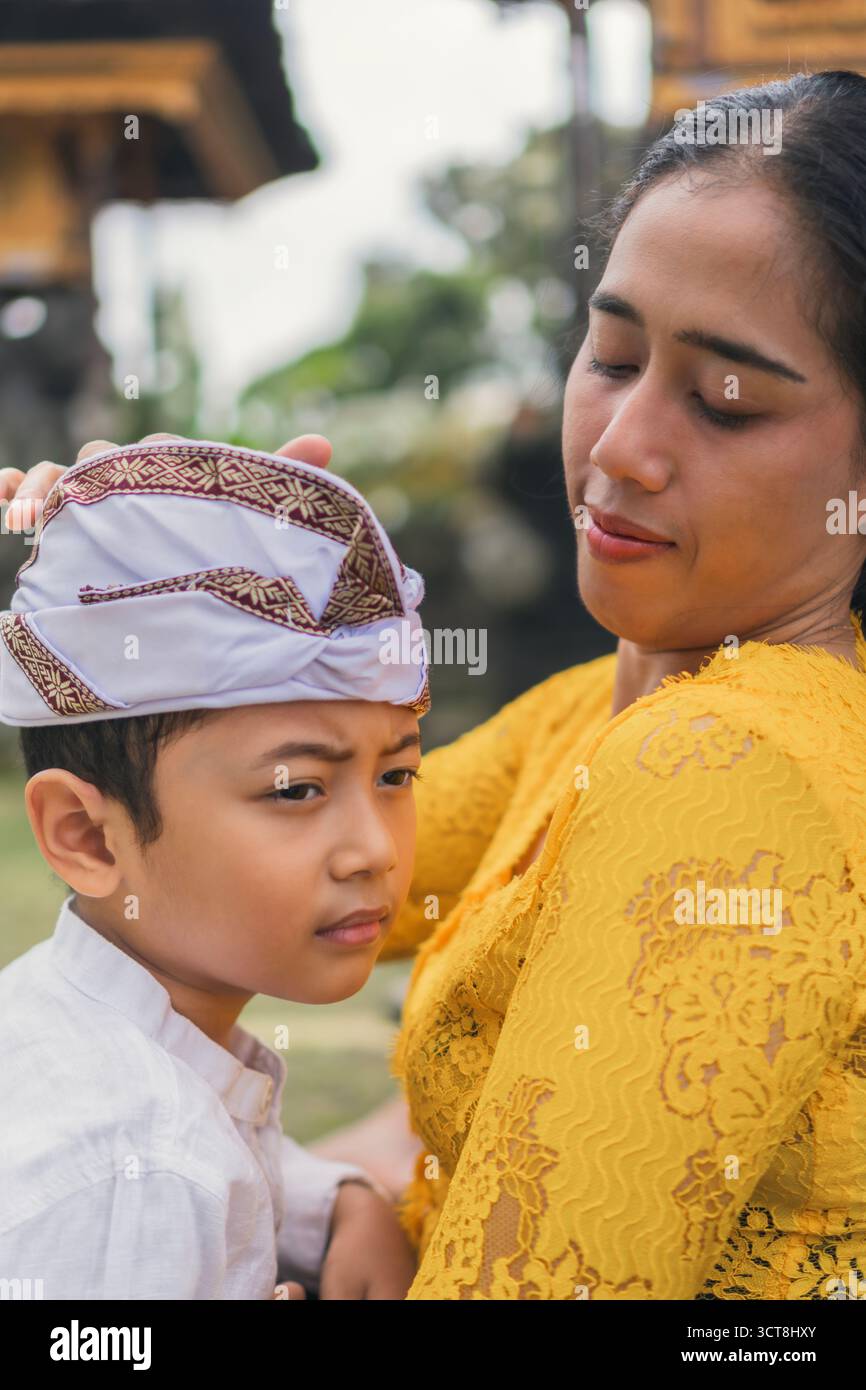 Una madre balinese che tiene in braccio il bambino mentre regola il copricapo udeng durante le preghiere Galungan al tempio. Foto Stock