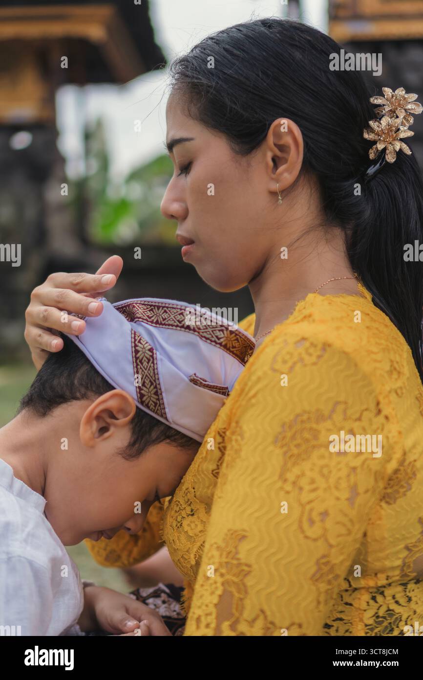 Una madre che tiene delicatamente la testa del bambino durante le preghiere Galungan in un tempio balinese, mostrando affetto e cura. Foto Stock