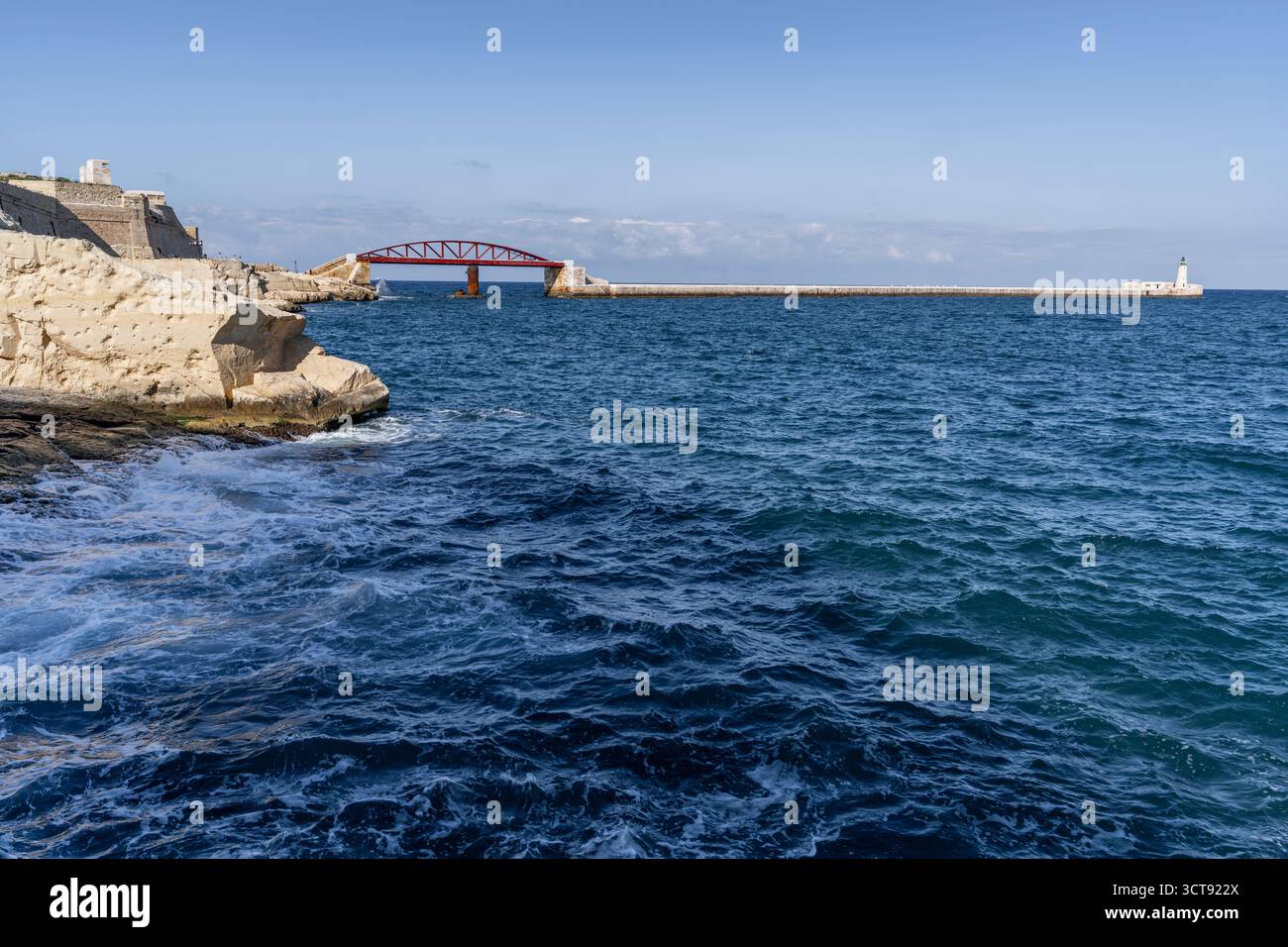 Onde schiumose che colpiscono la costa rocciosa, fortificazioni storiche e iconico ponte rosso sull'orizzonte blu del mare. Foto Stock