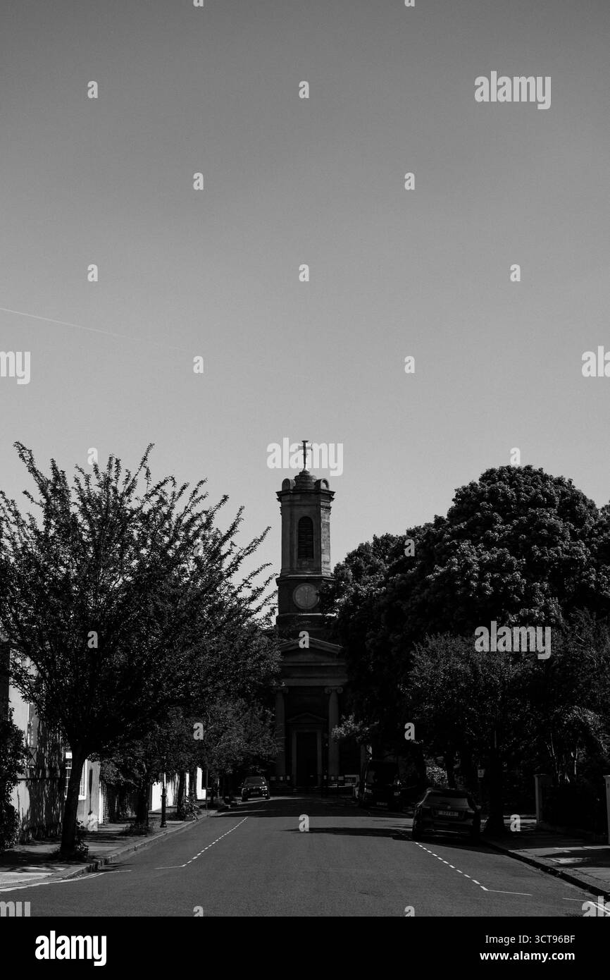 Chiswick, Londra – 2025.04.28 – Una fotografia in bianco e nero di una torre di una chiesa che si erge sopra le cime degli alberi alla fine di una tranquilla strada residenziale, Foto Stock