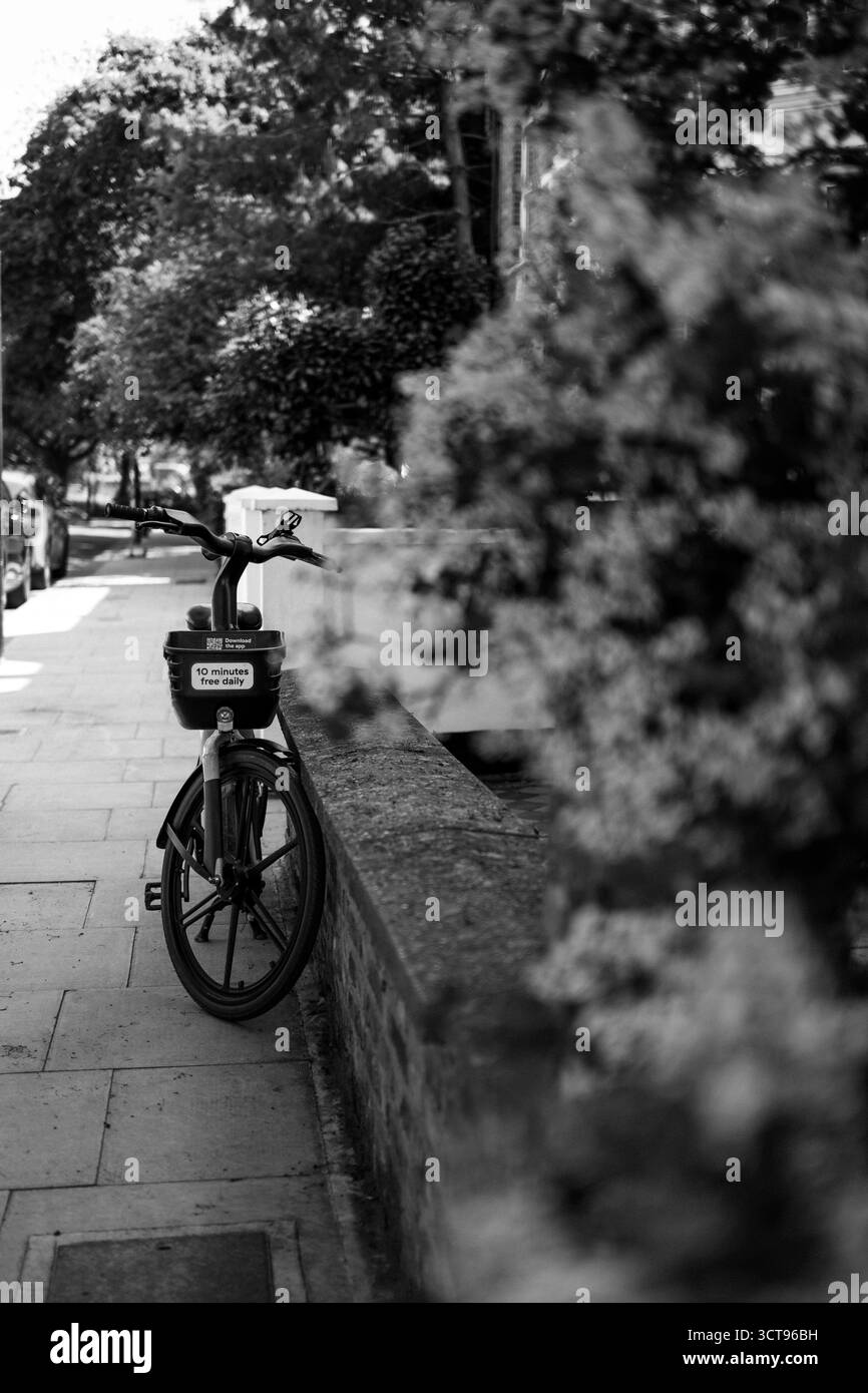 Chiswick, Londra – 2025.04.28 – foto in bianco e nero di una bicicletta a noleggio pubblica parcheggiata in una tranquilla strada residenziale accanto a un muro di mattoni coperto Foto Stock