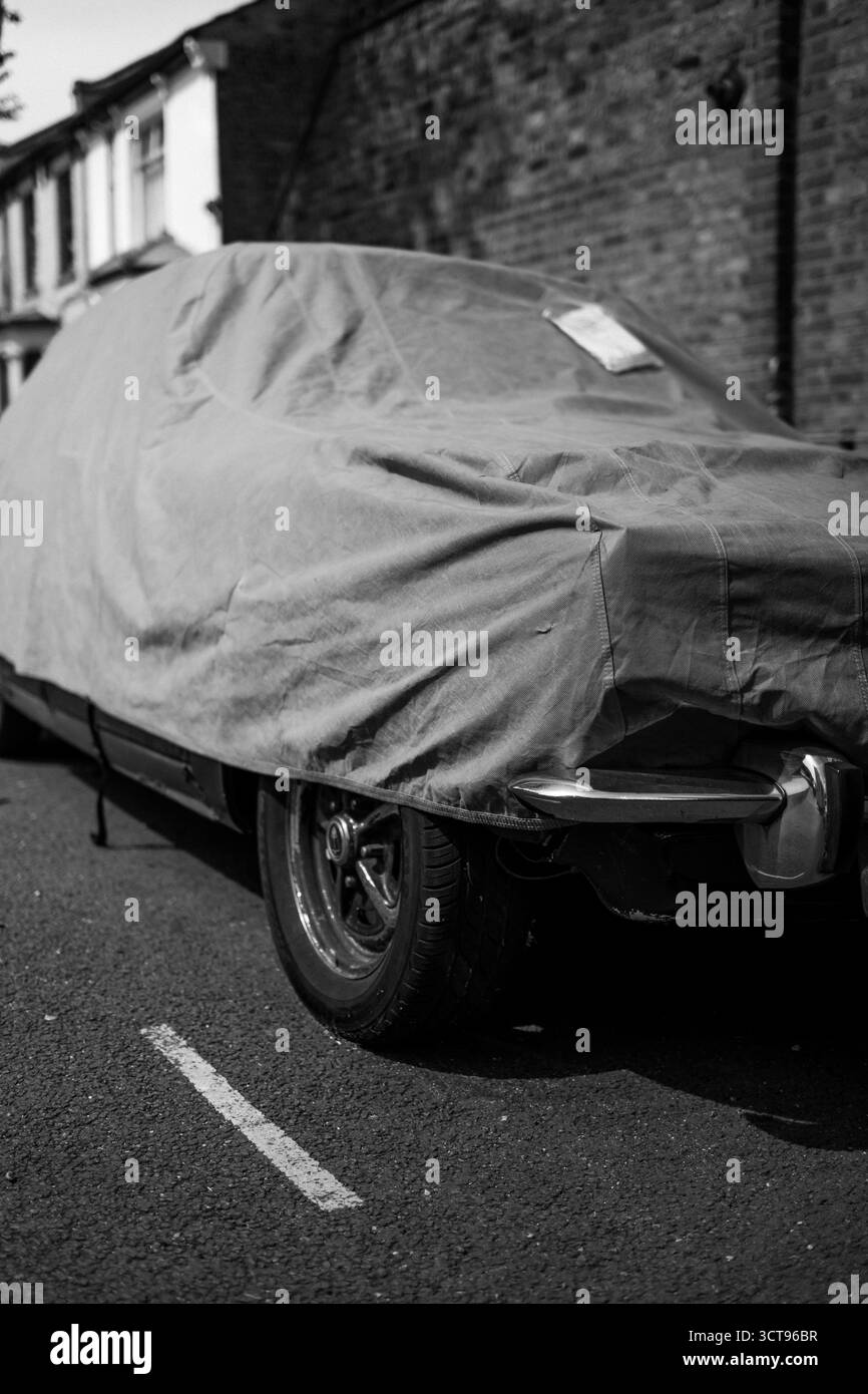 Chiswick, Londra – 2025.04.28 – Un'auto d'epoca coperta da un telo protettivo è parcheggiata lungo una tranquilla strada residenziale, con pareti in mattoni e terrazze Foto Stock