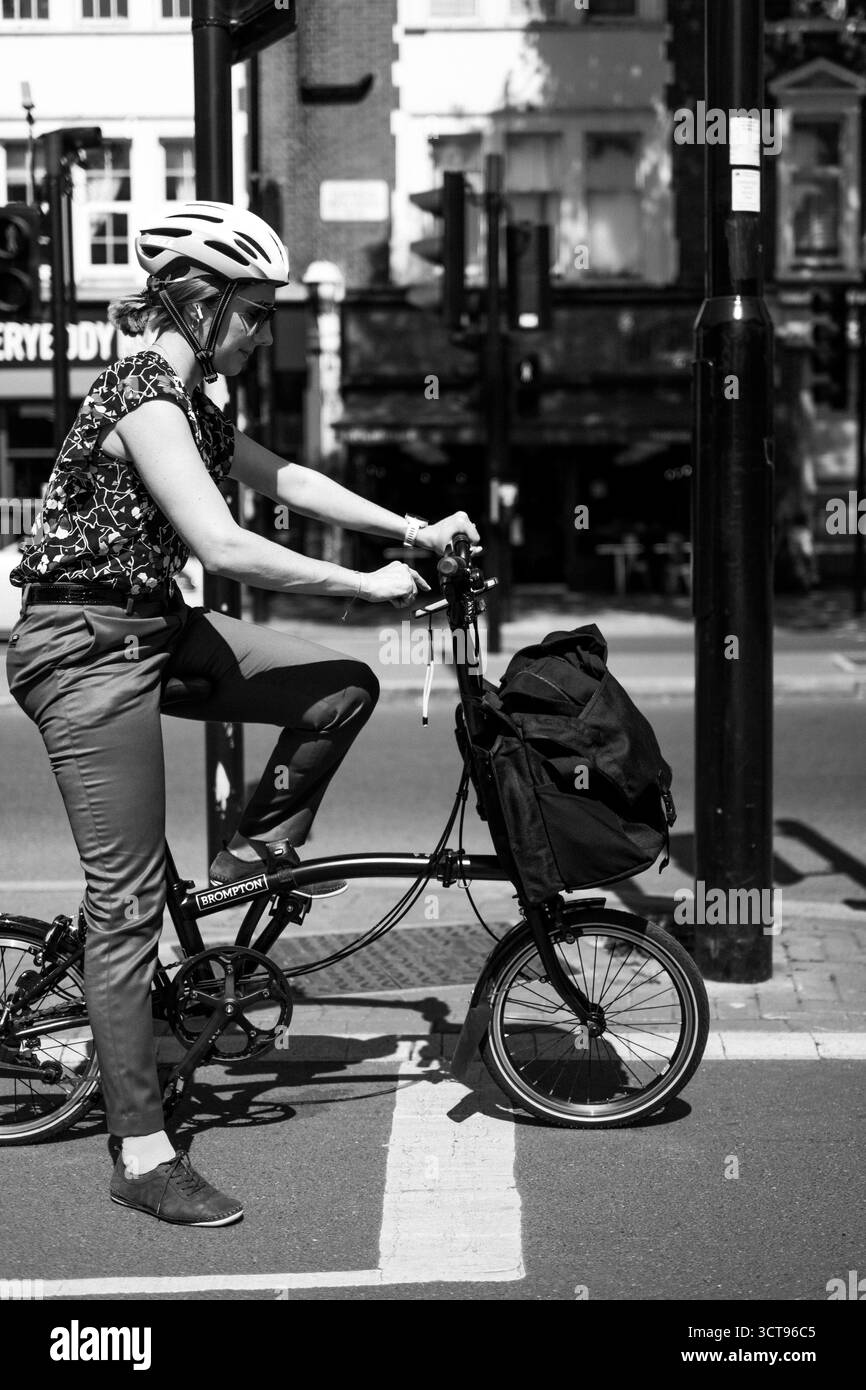 Chiswick, Londra – 2025.04.28 – Un pendolare che indossa un casco si ferma al semaforo su una bici pieghevole Brompton, tenendo una borsa sul manubrio Foto Stock