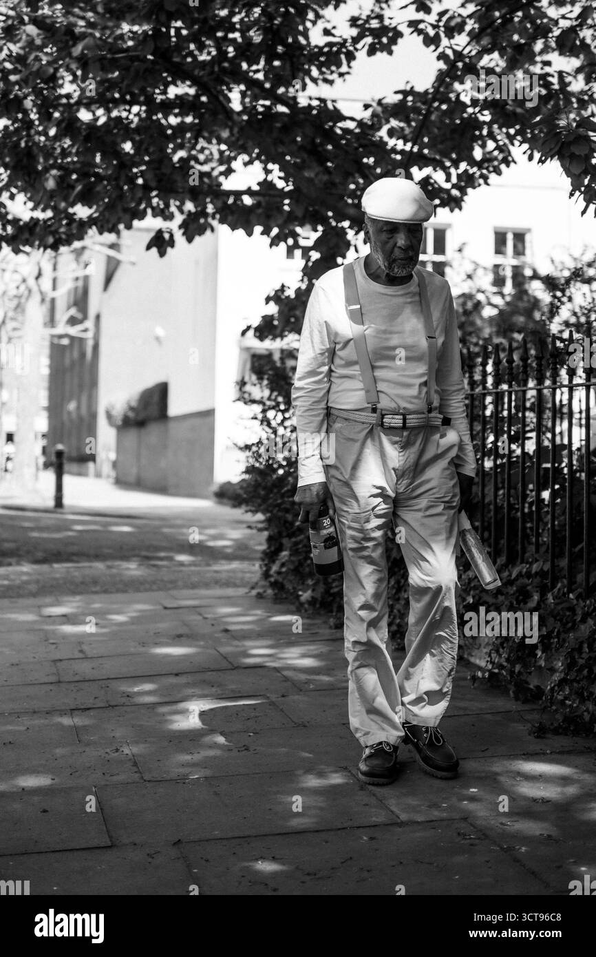 Chiswick, Londra – 2025.04.28 – foto candida in bianco e nero di un uomo anziano con abiti leggeri e bretelle che cammina lungo un sentiero ombreggiato di strada Foto Stock