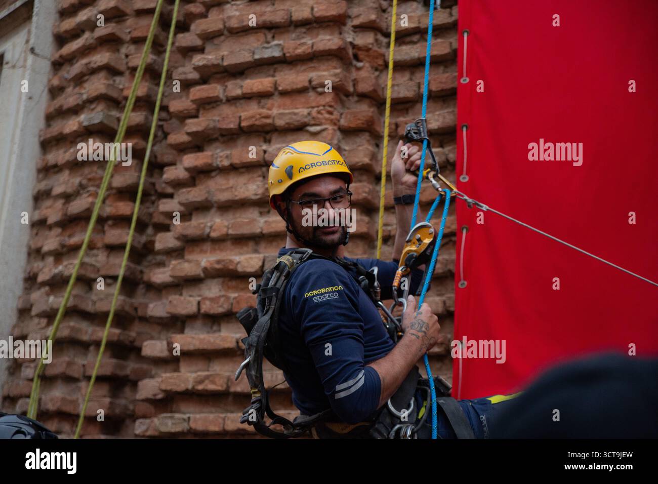 Un tecnico di fune di Acrobatica si vede salire attivamente sulla tela rossa installata, spostandosi in una posizione strategica per assistere e garantire la sicurezza Foto Stock