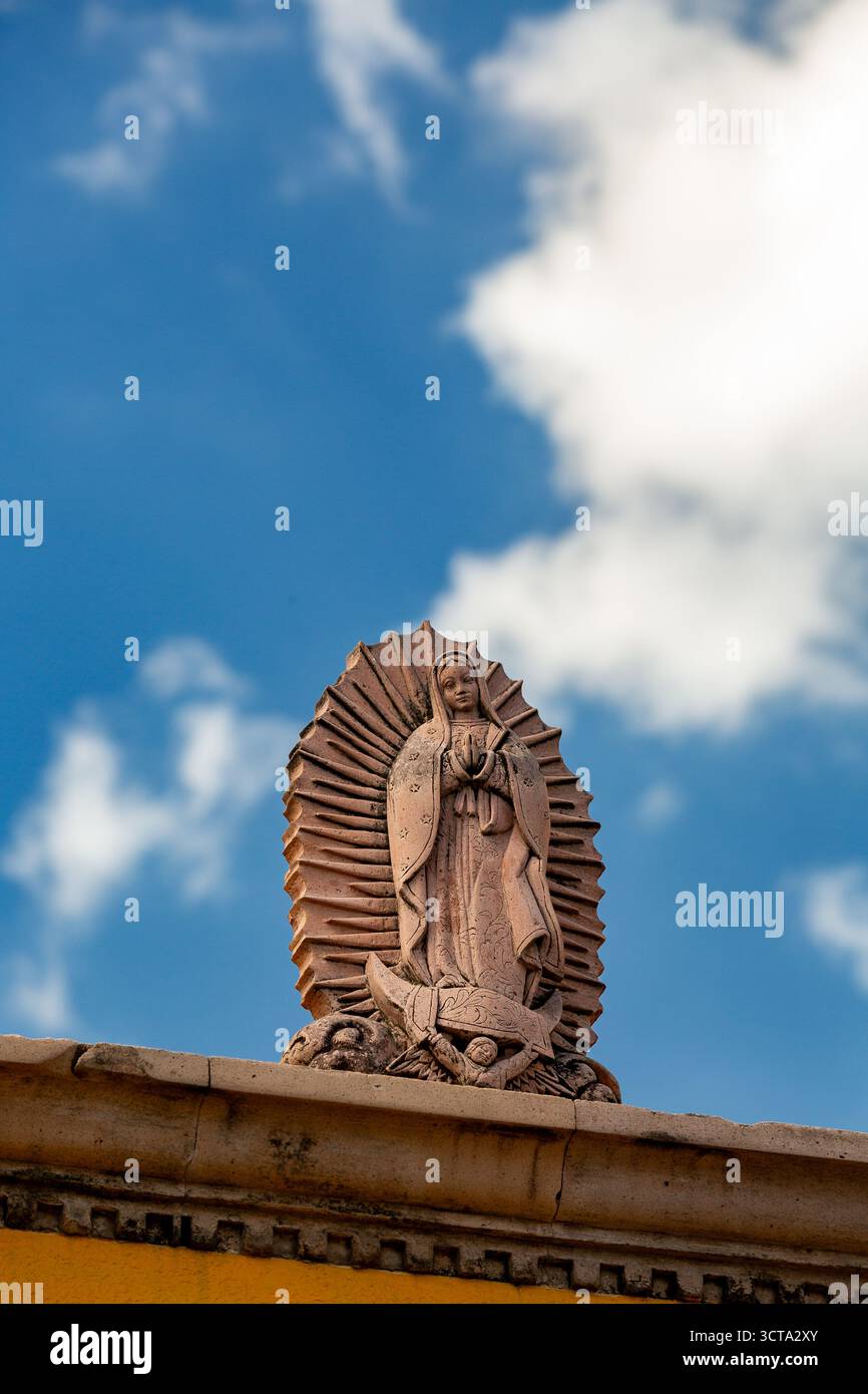 Una statua della Vergine Maria si erge in cima a un edificio, le sue mani si aggrappano in preghiera su uno sfondo di cielo blu e soffici nuvole bianche. Foto Stock