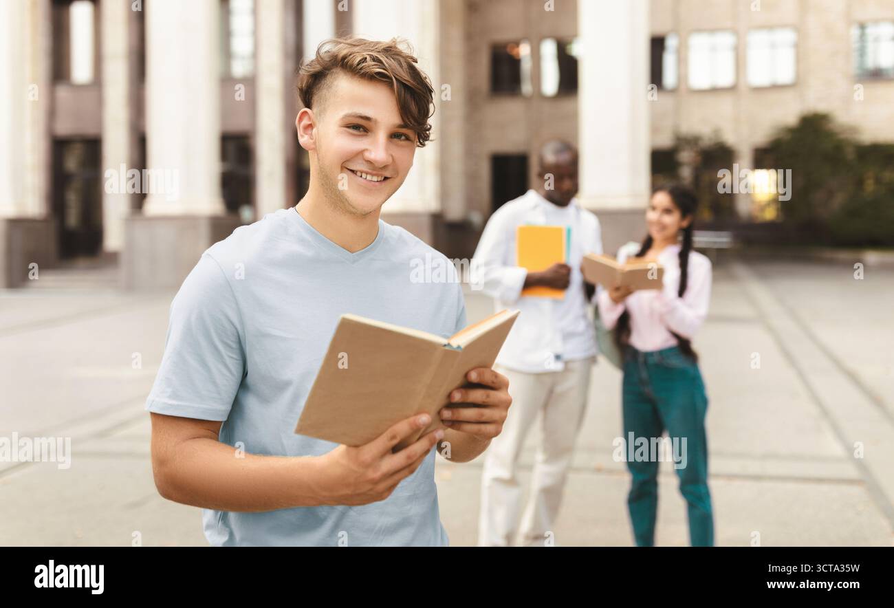 Corsi di laurea all'estero. Ritratto di felice studente maschio con libro in posa all'aperto al campus con i suoi compagni di classe Foto Stock