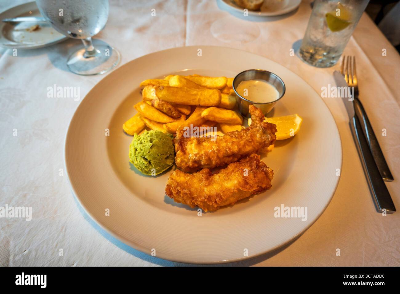 Classico fish and chips con merluzzo pastella dorato, patatine fritte croccanti, vivaci piselli e spicchi di limone, serviti con un lato di cremosa salsa tartare Foto Stock