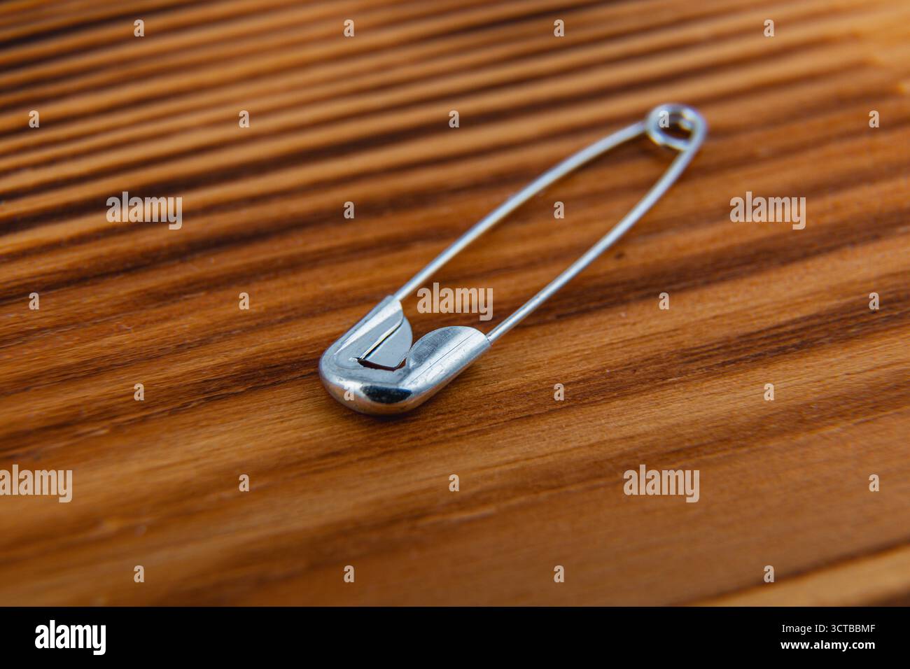 Una spilla di sicurezza color argento si trova su una superficie in legno testurizzato, evidenziata dalla luce naturale, che mostra la sua finitura lucida e il design semplice contro il caldo a. Foto Stock