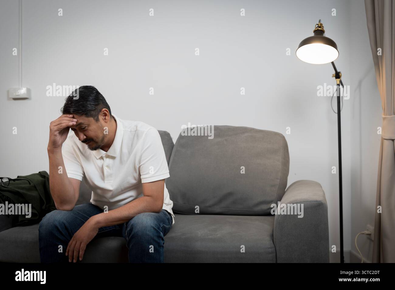 Un uomo asiatico premuroso, a metà degli anni '40, siede da solo sul suo divano in una stanza poco illuminata, tenendo la testa in profondità. Questa immagine rappresenta la guarigione mentale maschile Foto Stock