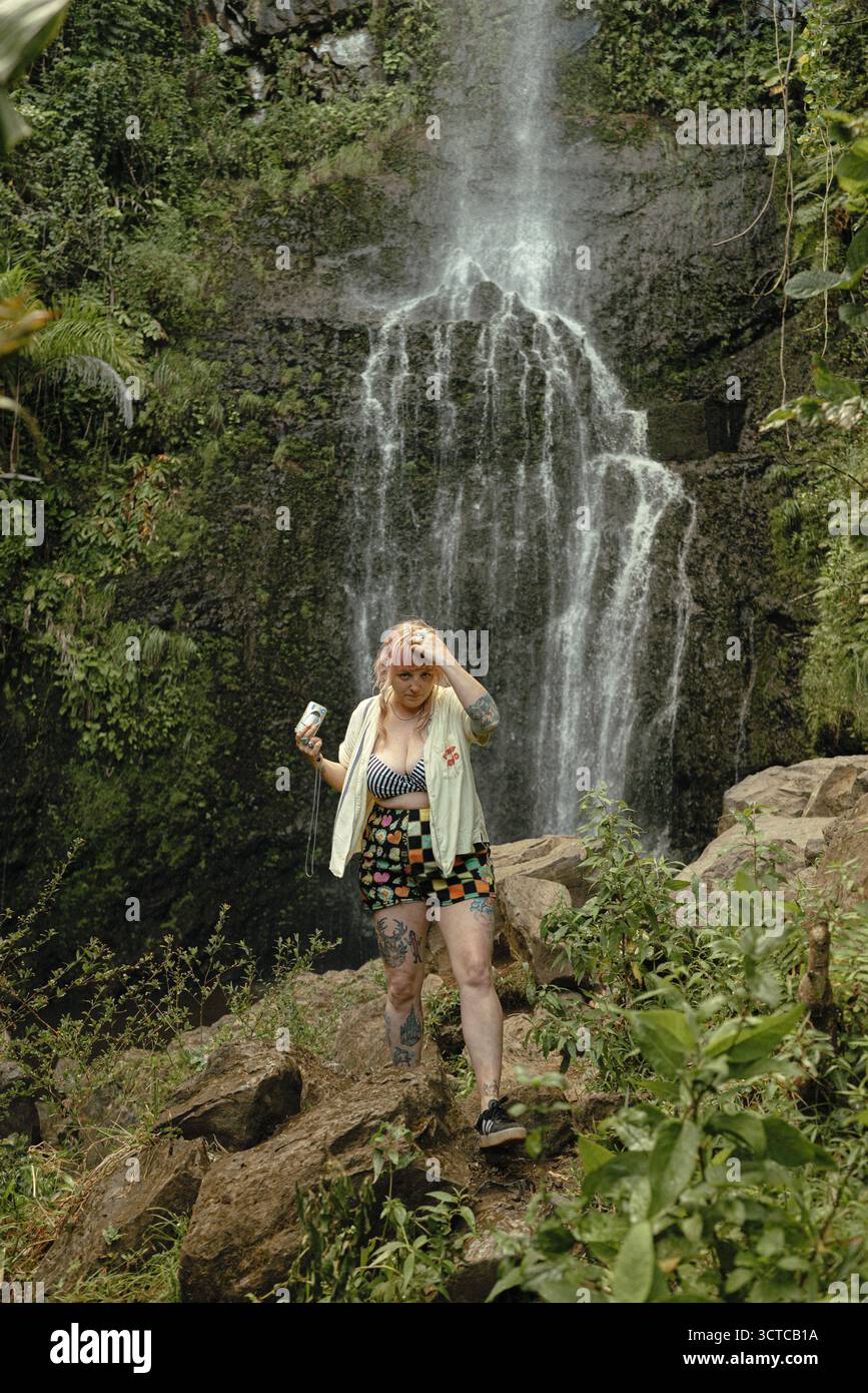 Una donna tatuata si avvicina alla nebbiosa cascata di Maui con una macchina fotografica Foto Stock