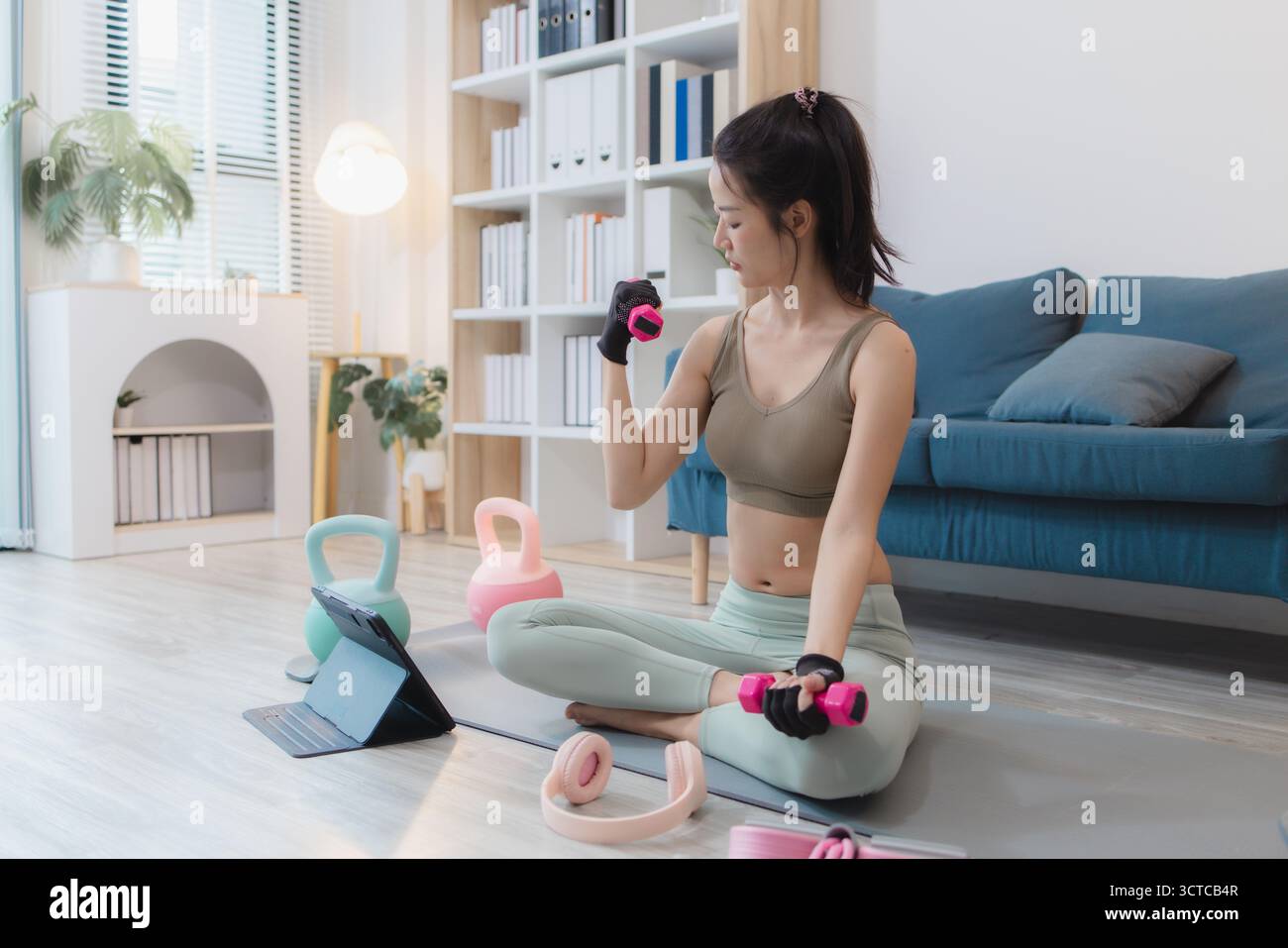 Una giovane donna si sta esercitando a casa, praticando yoga e allenamento di forza con manubri e kettlebell su un tappetino. Segue la sua routine di allenamento wi Foto Stock