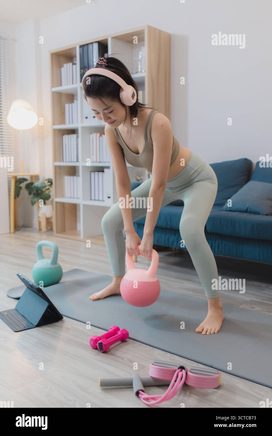 Una giovane donna si sta esercitando a casa, praticando yoga e allenamento di forza con manubri e kettlebell su un tappetino. Segue la sua routine di allenamento wi Foto Stock