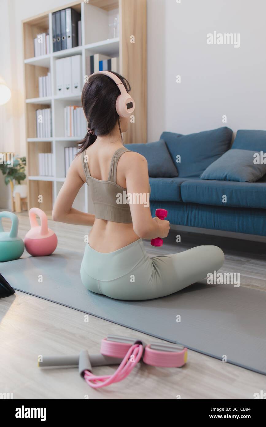 Una giovane donna si sta esercitando a casa, praticando yoga e allenamento di forza con manubri e kettlebell su un tappetino. Segue la sua routine di allenamento wi Foto Stock
