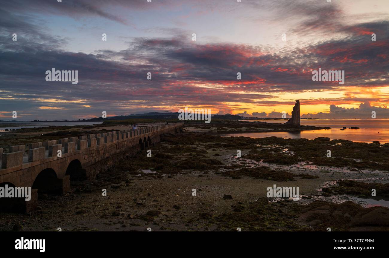 Il tramonto si illumina dietro il vecchio faro costiero e le onde si infrangono. Foto Stock