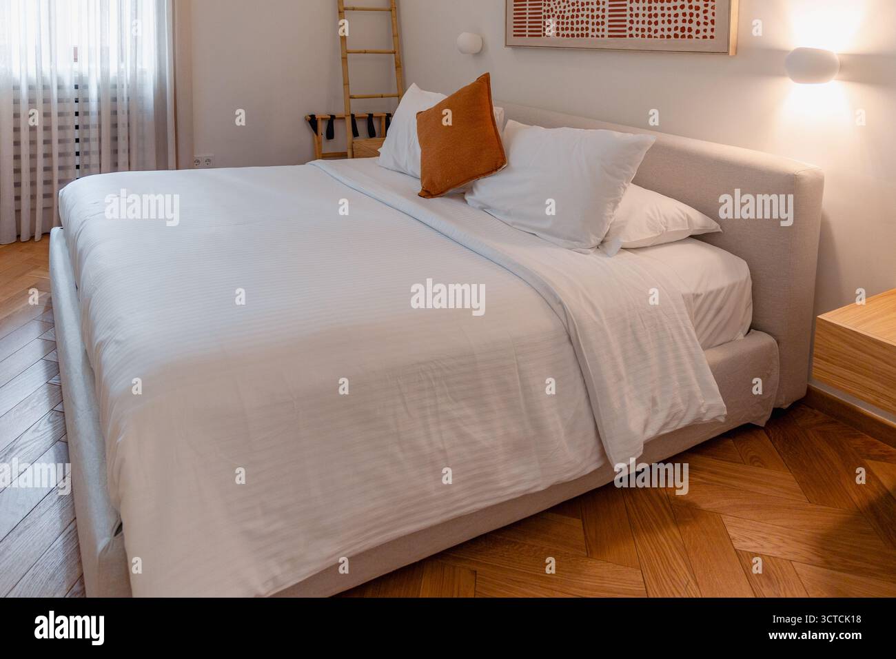 Una comoda camera da letto con fresche lenzuola bianche su un letto moderno. Presenta pavimento in parquet a spina di pesce e luce calda e soffusa da una applique Foto Stock