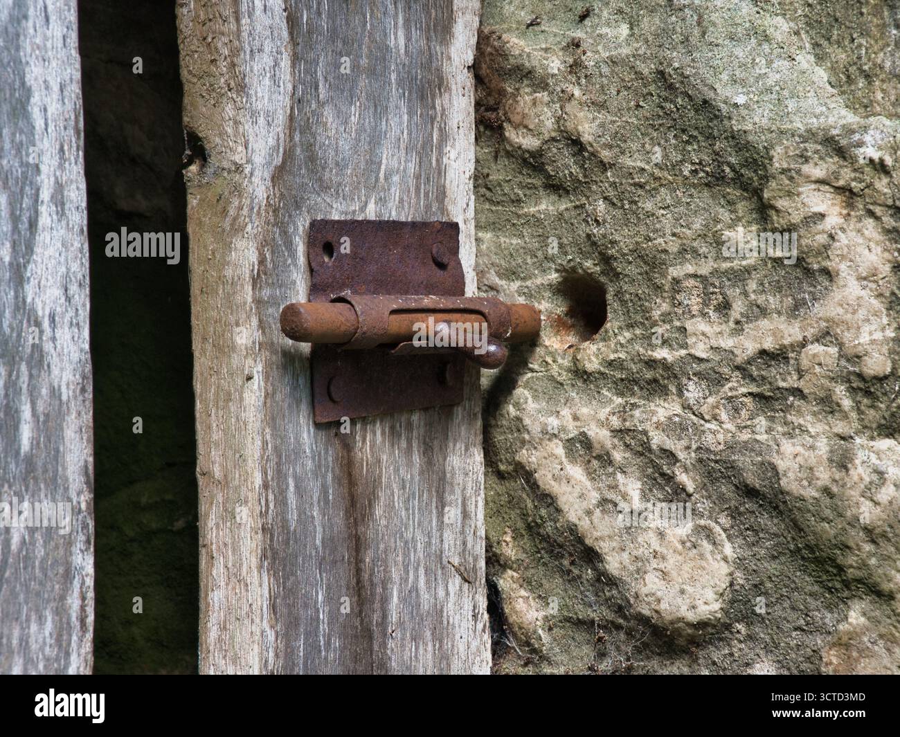 Serratura in metallo ossidato su legno e pietra antichi - primo piano di un bullone scorrevole pesantemente arrugginito contro tavole di legno intemperie e una struttura ruvida della parete in pietra. Foto Stock