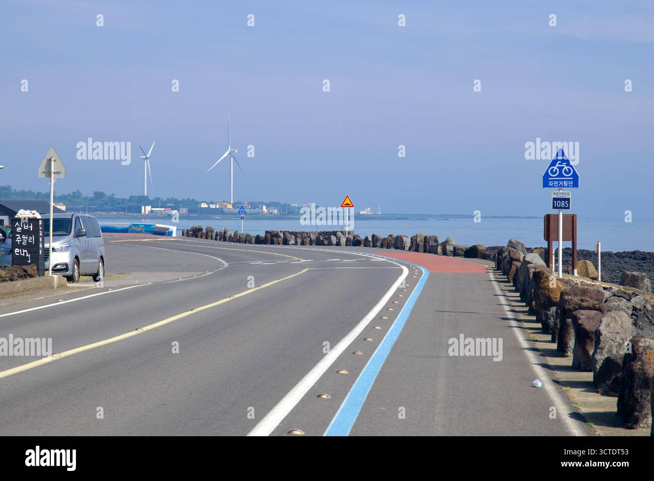 Un tratto curvo della strada costiera di Handong mostra la pista ciclabile blu sopra le rocce vulcaniche, con turbine eoliche che girano in lontananza lungo il Jeju sh Foto Stock