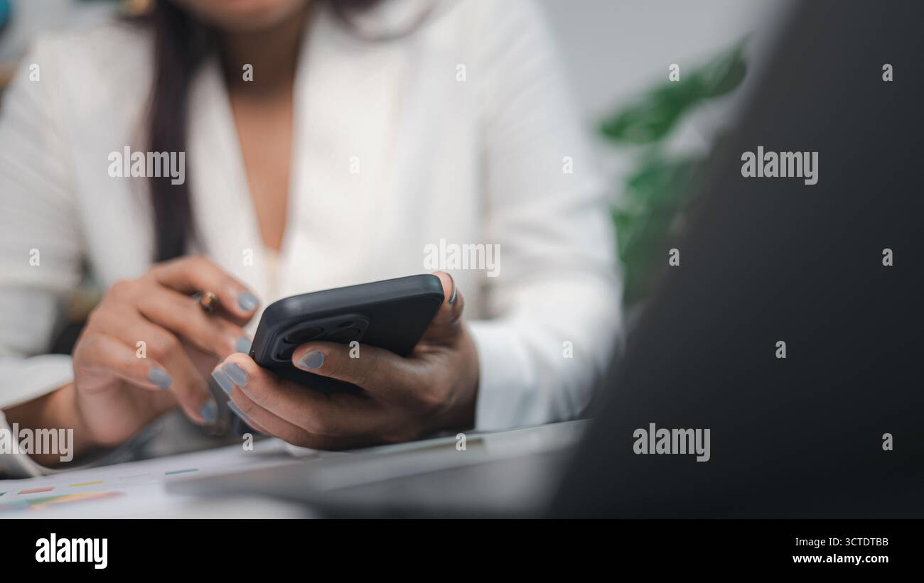 Donna d'affari che tiene in mano lo smartphone mentre lavora con carte finanziarie e laptop sulla scrivania dell'ufficio. Concetto di comunicazione digitale, tecnologia, online f Foto Stock