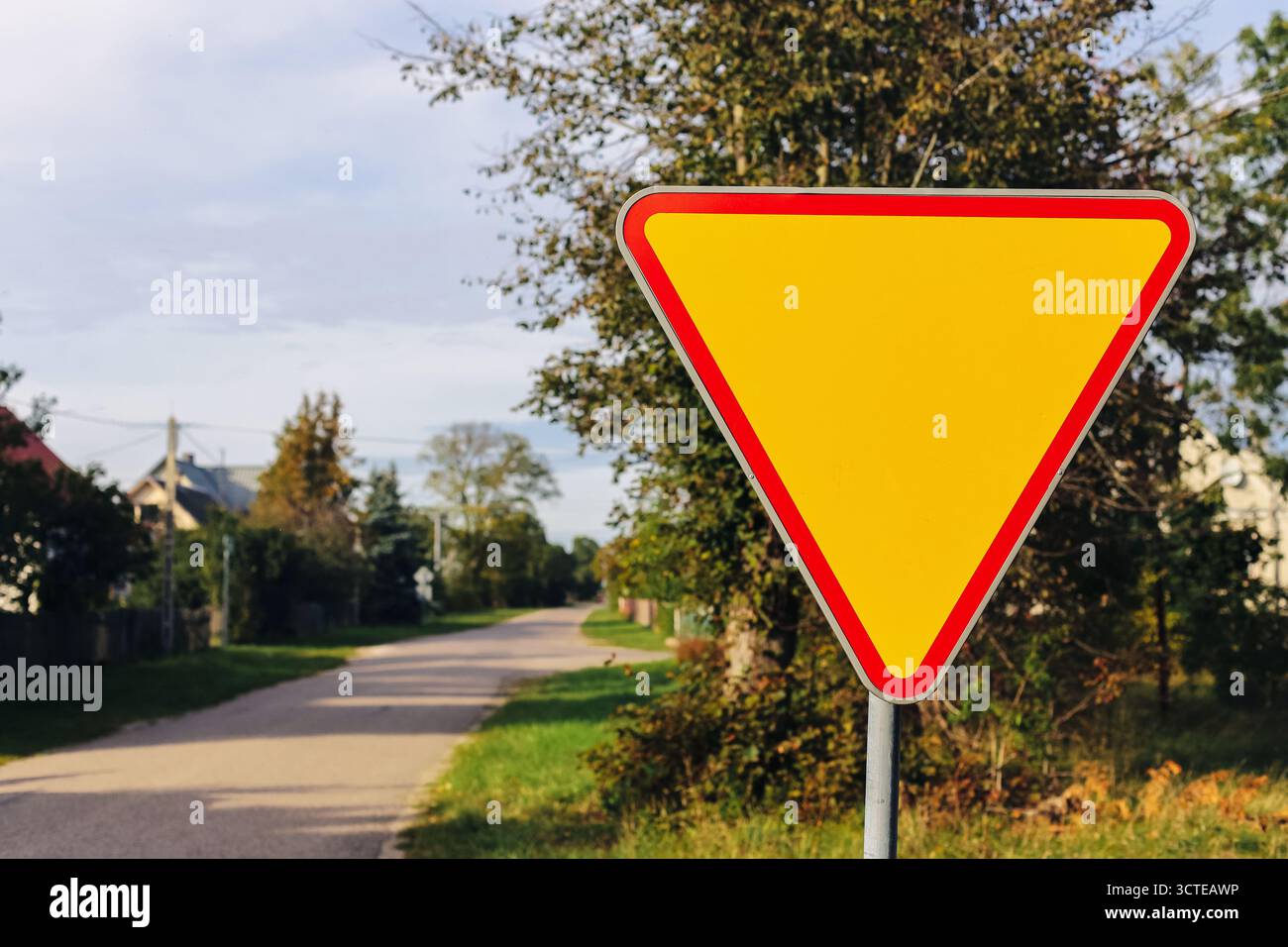 Cartello triangolare sulla strada di campagna con cielo limpido. Simbolo del controllo del traffico e della sicurezza stradale nelle zone rurali con alberi e case. Foto Stock