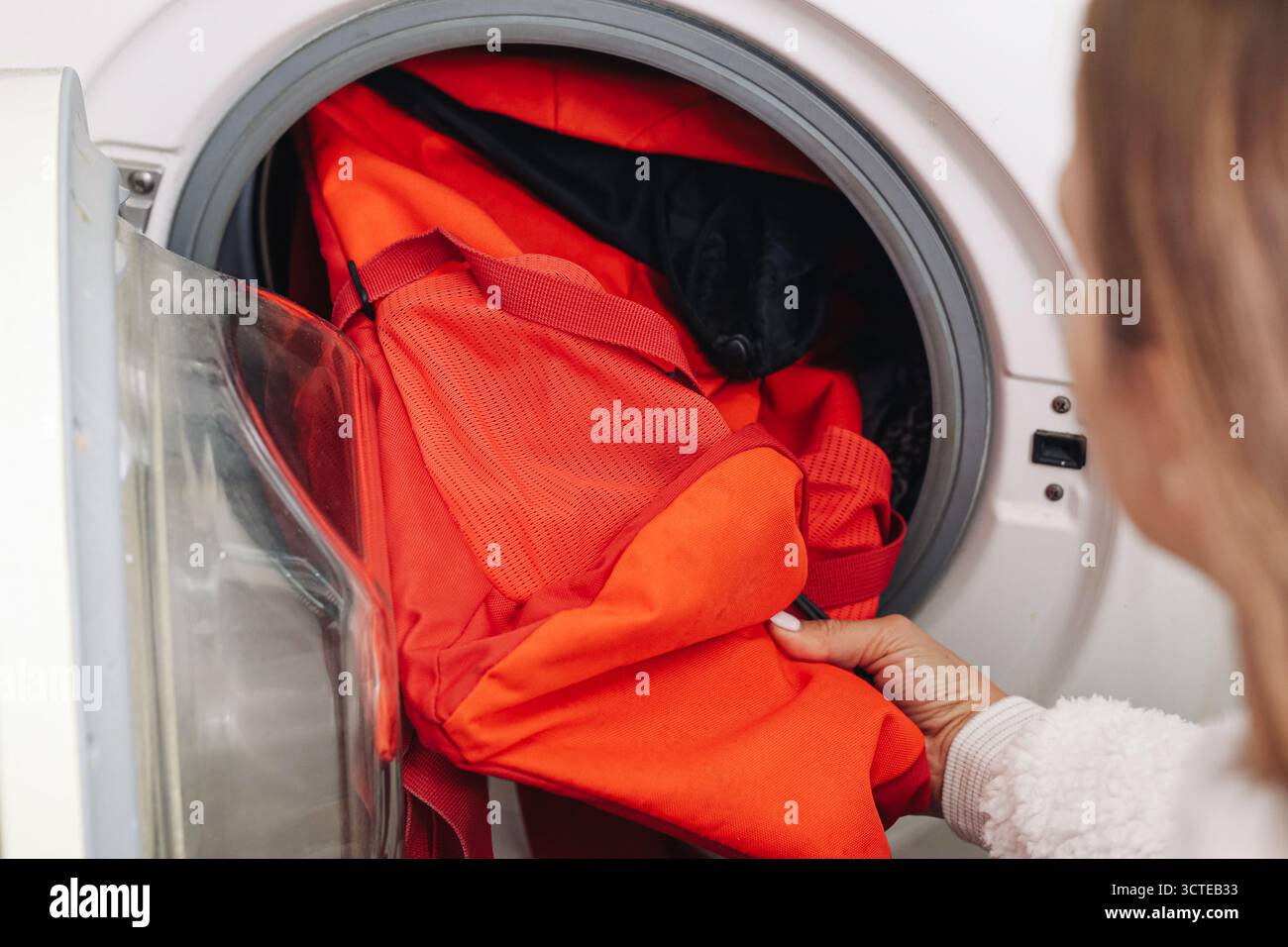 Donna che mette lo zaino rosso nel tamburo della lavatrice. Sacchetto di tessuto per la pulizia a casa. Zaino da viaggio per la cura del bucato e il lavaggio per l'igiene. Foto Stock