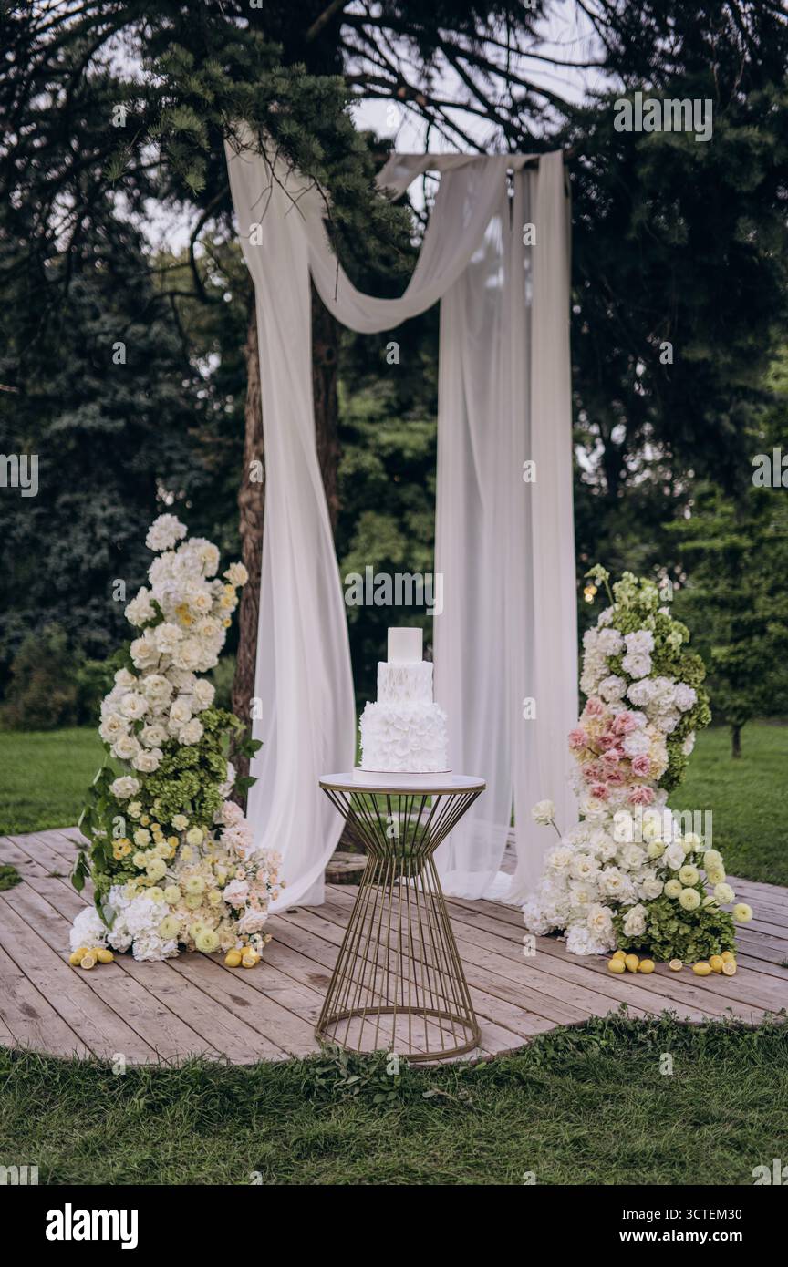 Una pittoresca location per matrimoni presenta un'elegante torta su un piedistallo unico, circondato da incantevoli composizioni floreali e morbidi tendaggi, perfetto per un medico Foto Stock