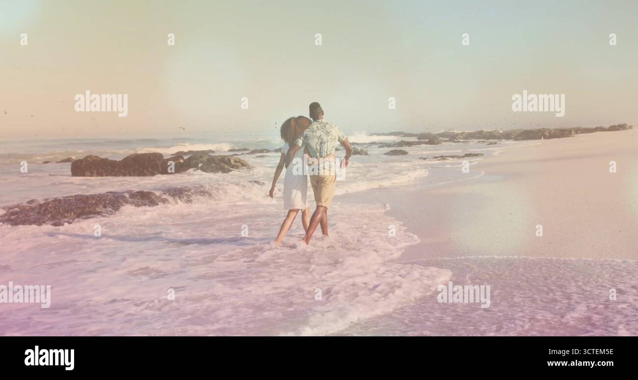 Una coppia afro-americana a braccia nude per fare surf sulla spiaggia sabbiosa, con onde e rocce scure Foto Stock