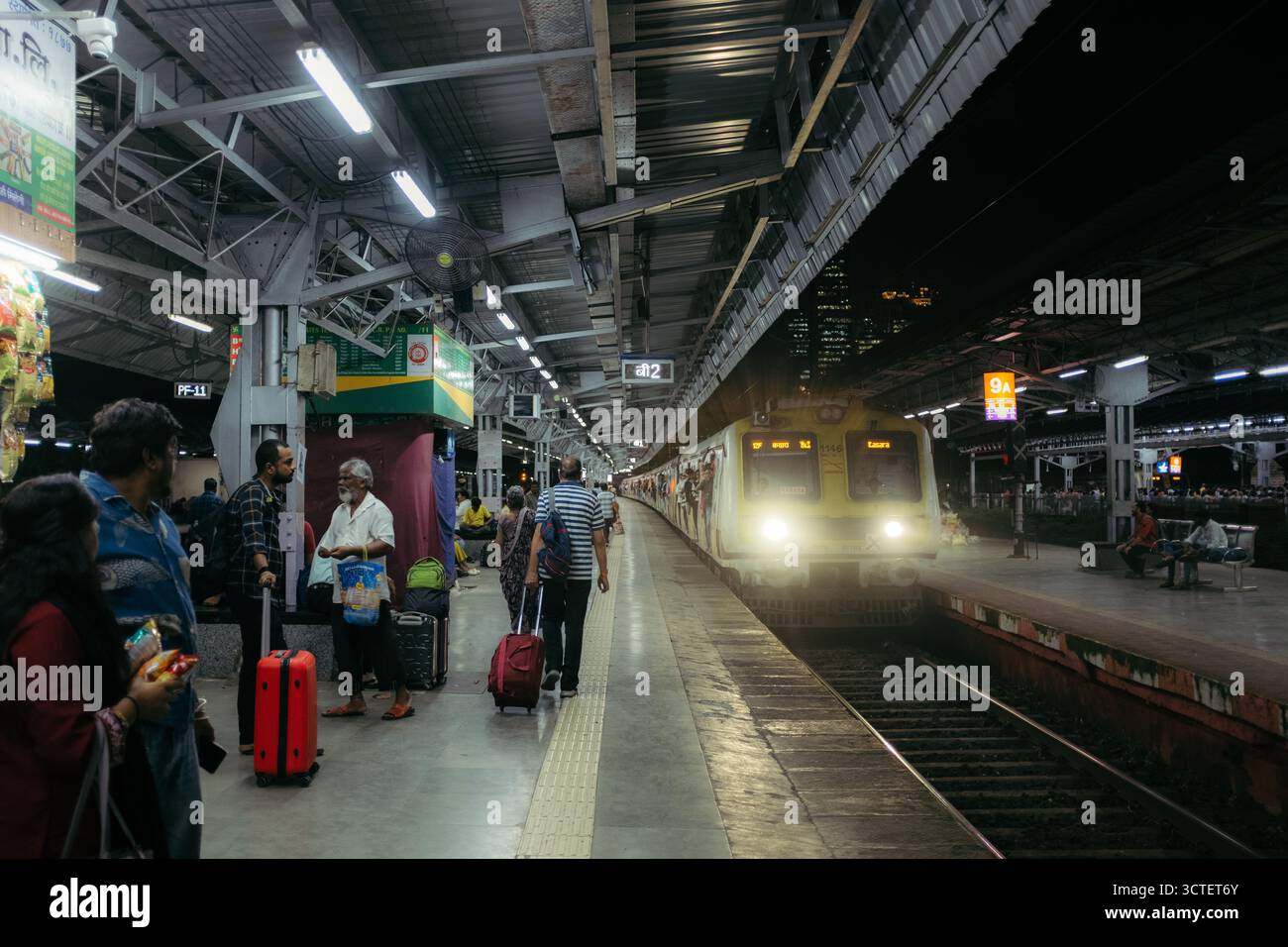 Mumbai, India - 23 settembre 2025: Veduta di una vivace piattaforma ferroviaria dove ti aspetta un treno illuminato che getta lunghe ombre tra pendolari e bagagli. Foto Stock