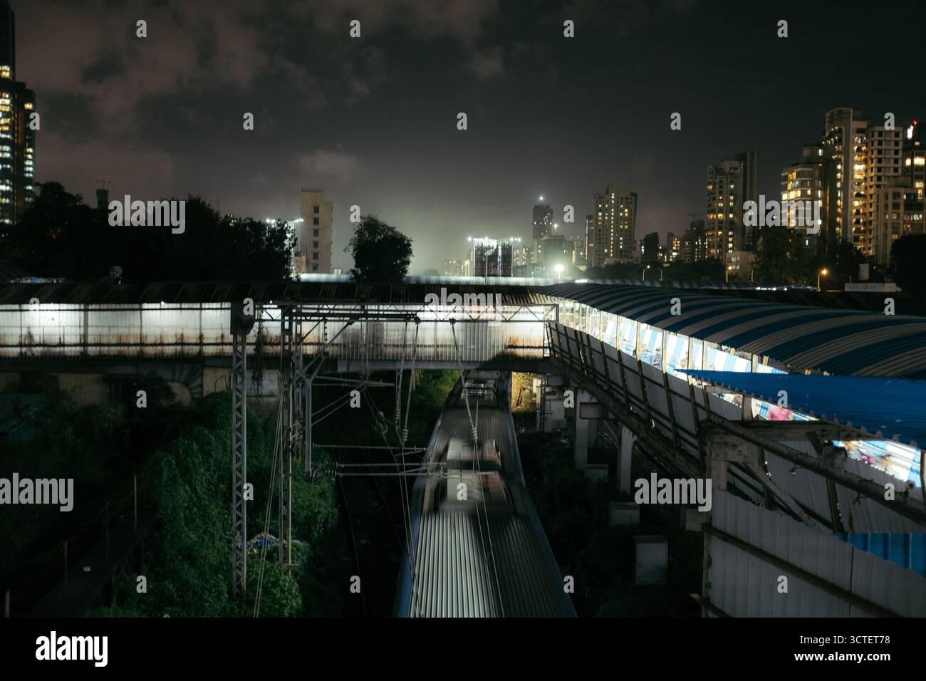 Mumbai, India - 23 settembre 2025: Vista di una stazione ferroviaria di notte, le luci della piattaforma proiettano un bagliore contro il cielo scuro e gli edifici lontani della città. Foto Stock