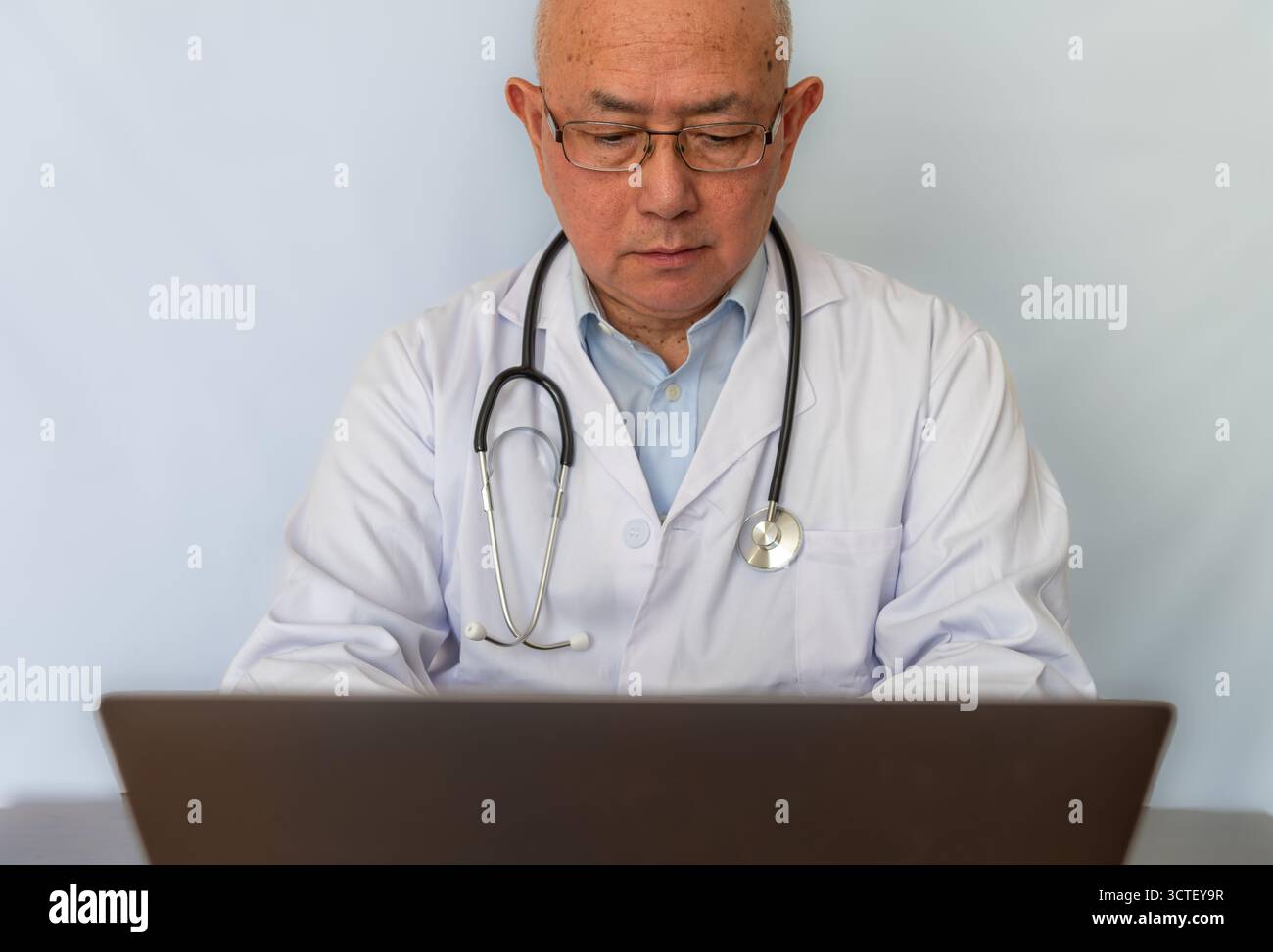 Un medico con un cappotto bianco con uno stetoscopio appeso al collo che lavora su un computer portatile su una scrivania. Foto Stock