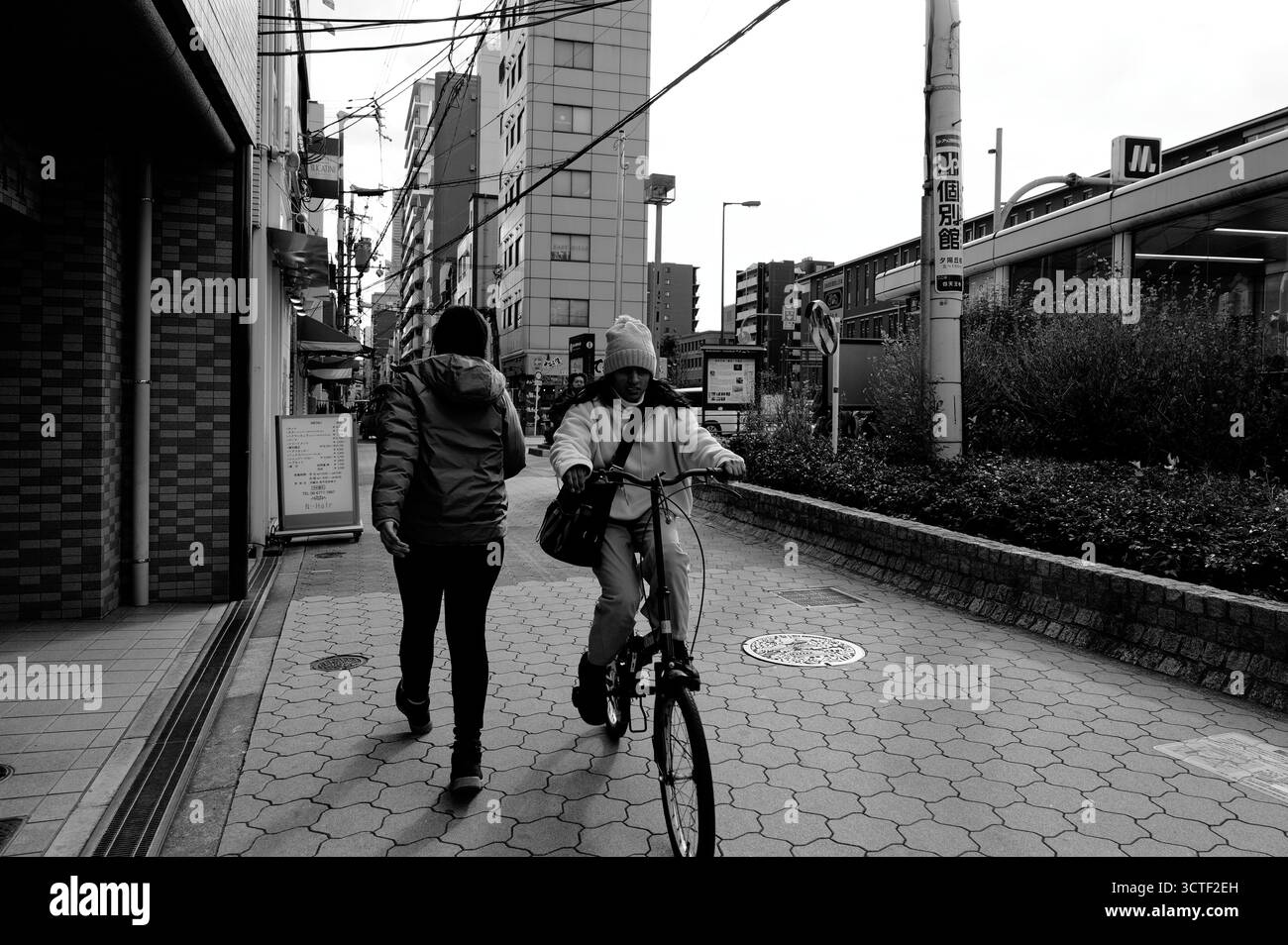 Un ciclista passa un pedonale in una tranquilla strada di Osaka, circondata da edifici di media altezza sotto un cielo nuvoloso Foto Stock