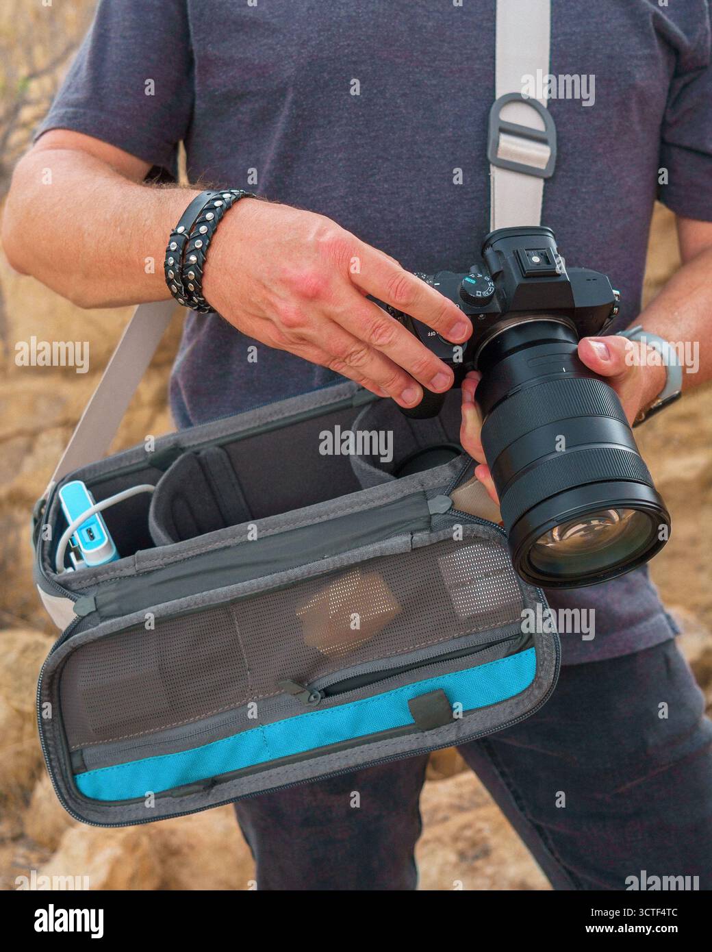 Uomo che tiene in mano la fotocamera per controllare le impostazioni con la borsa della fotocamera imbracata in primo piano nel deserto con sfondo roccioso. Copia spazio. Joshua Tree National Park, California Foto Stock