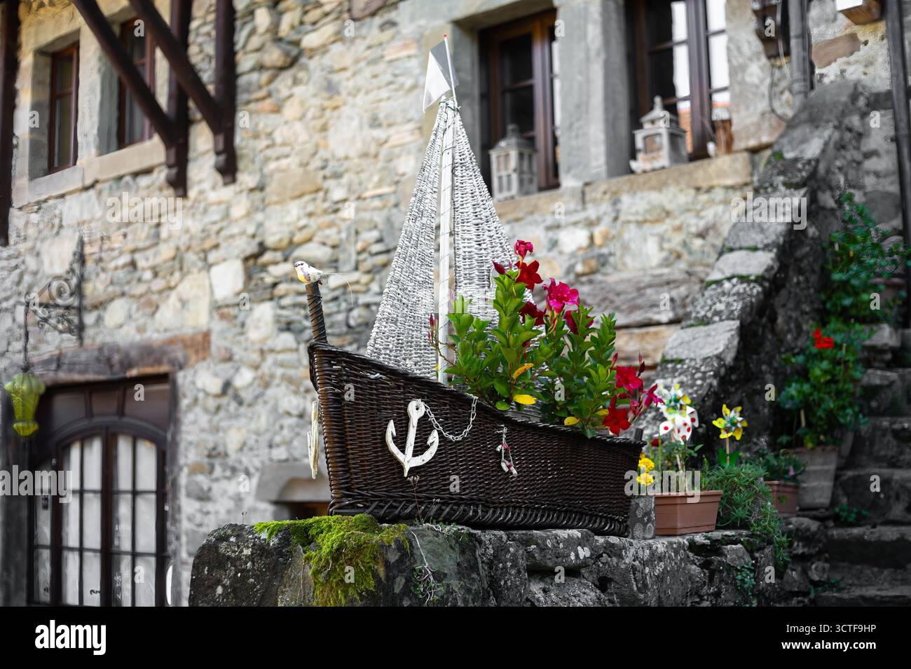 Yvoire, Francia - 25 settembre 2025: Un cesto decorativo in vimini a forma di barca, pieno di fiori vivaci, posto su un muro di pietra di fronte a Foto Stock