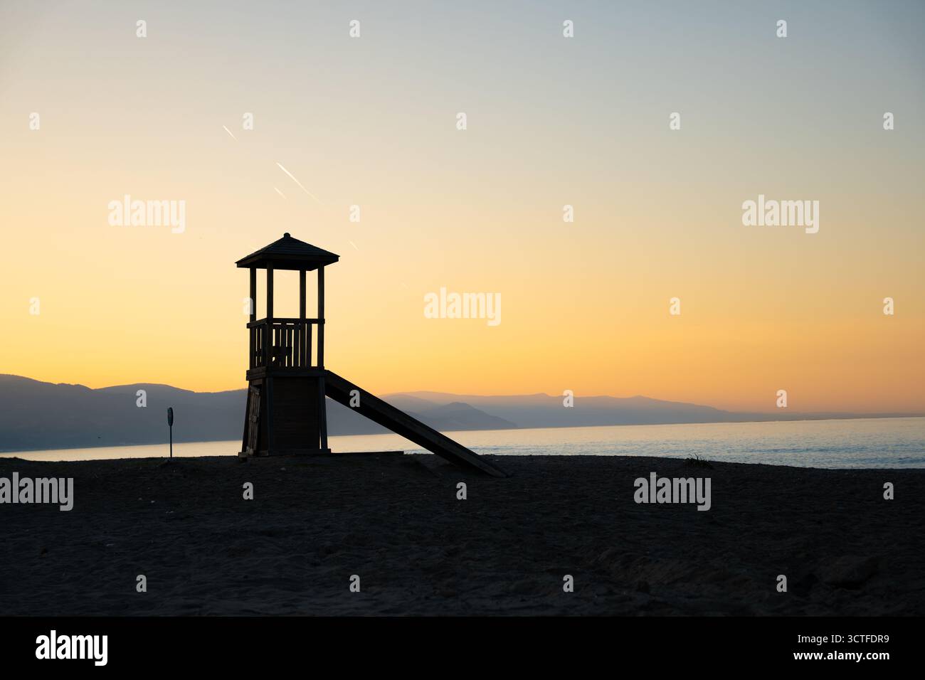 Torre bagnino sulla spiaggia al caldo tramonto, cielo arancione e montagne. Torre del bagnino in legno sagomato sul mare. Natura e sicurezza costiera Foto Stock