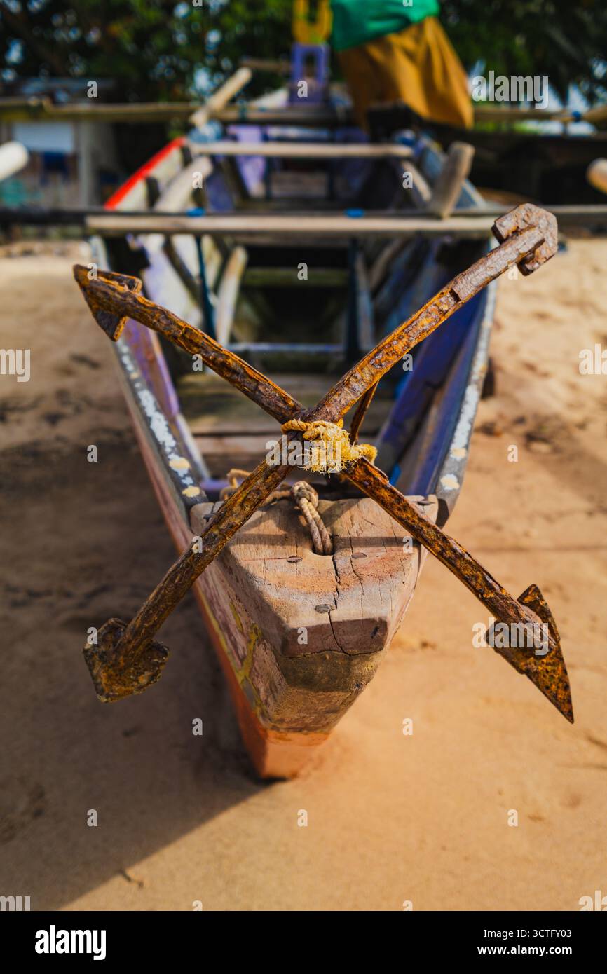 Primo piano di un'ancora in metallo arrugginito legata alla prua di una tradizionale barca da pesca in legno che poggia su una spiaggia sabbiosa. Foto Stock