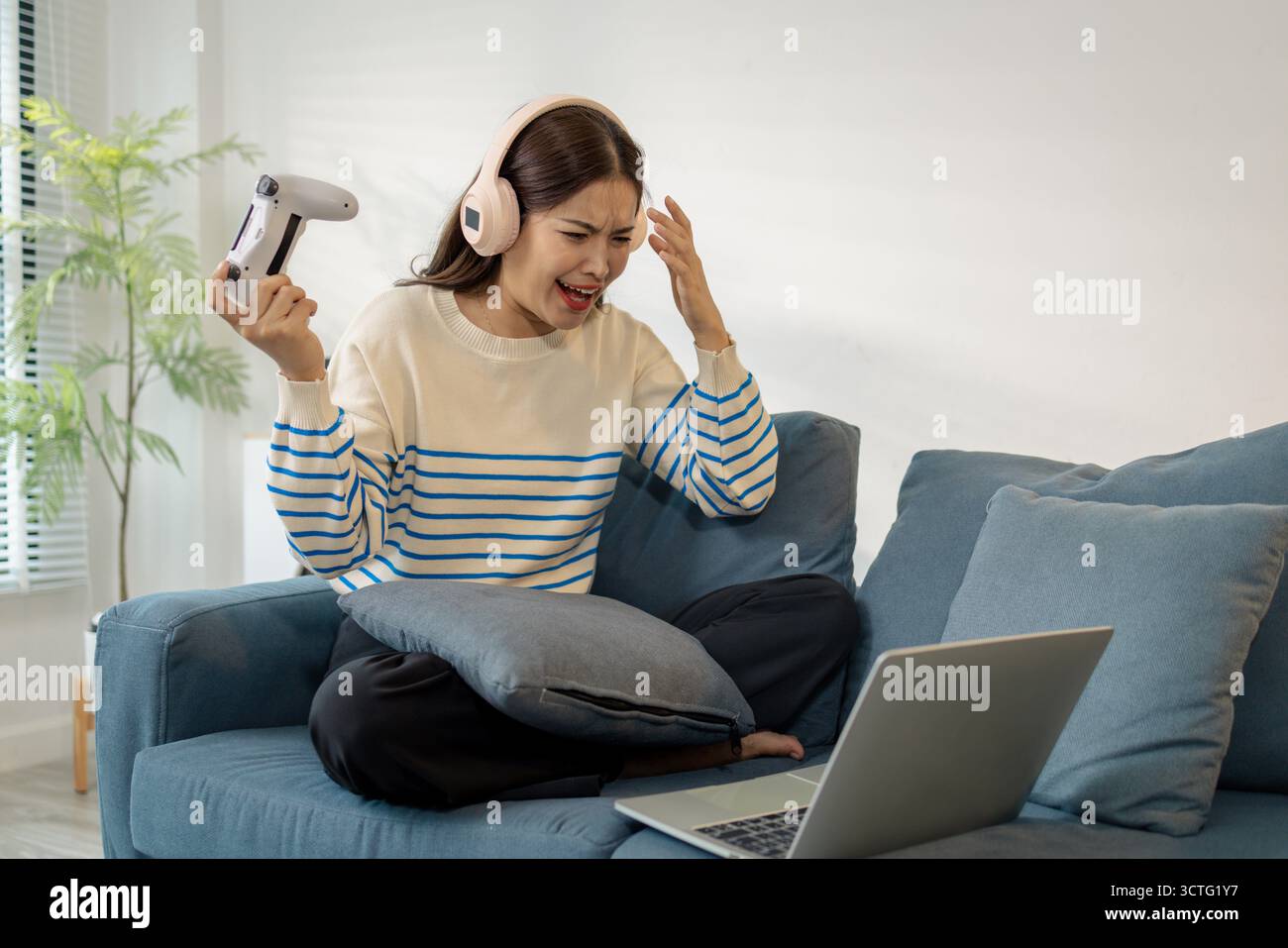Una giovane donna che indossa le cuffie sta giocando a un videogame sul suo notebook, tenendo in mano un controller con espressioni emotive di gioia, eccitazione e fruste Foto Stock
