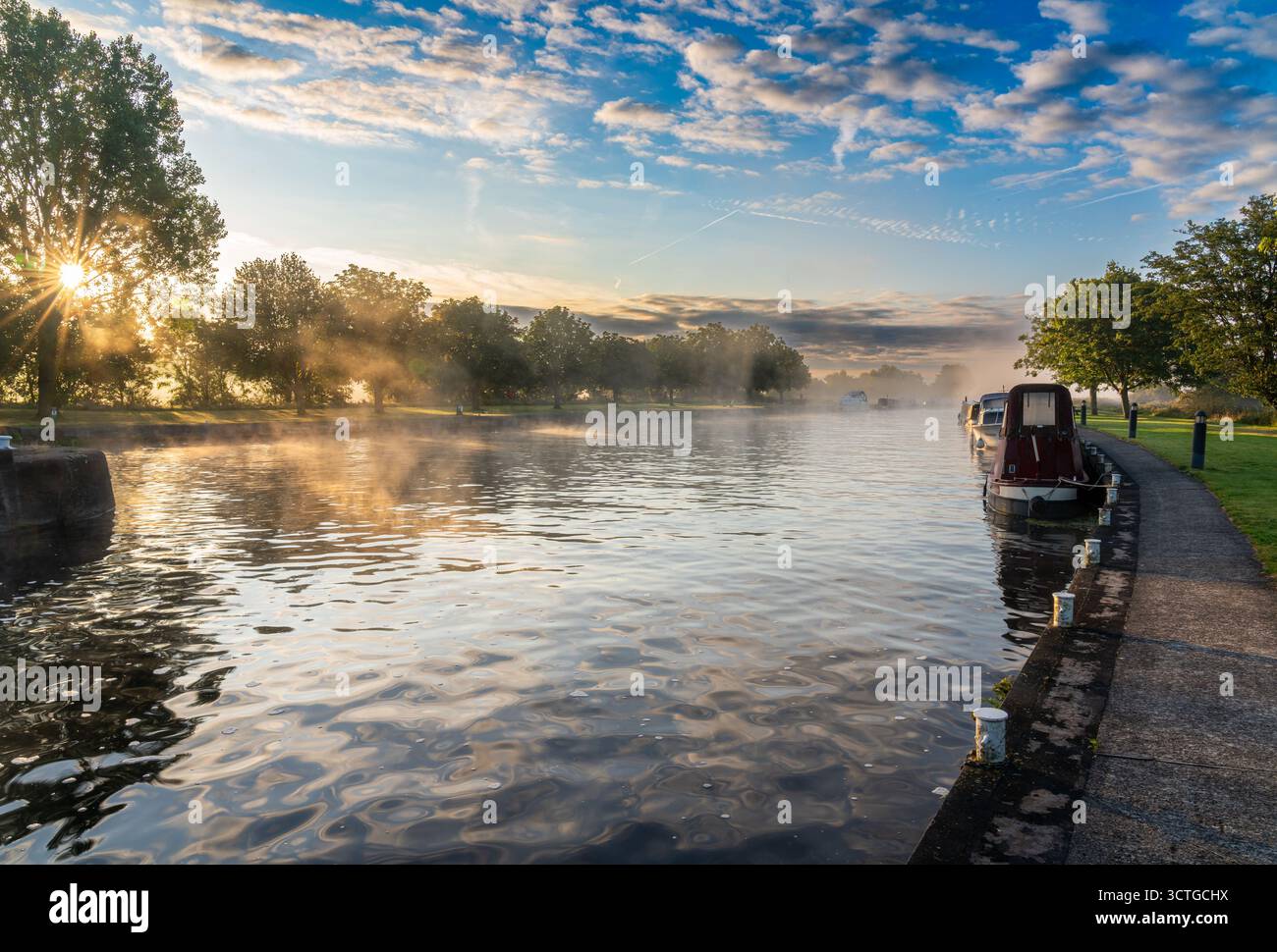 Nebbia mattutina sul canale calmo con barche ormeggiate e alberi illuminati dal sole all'alba Foto Stock