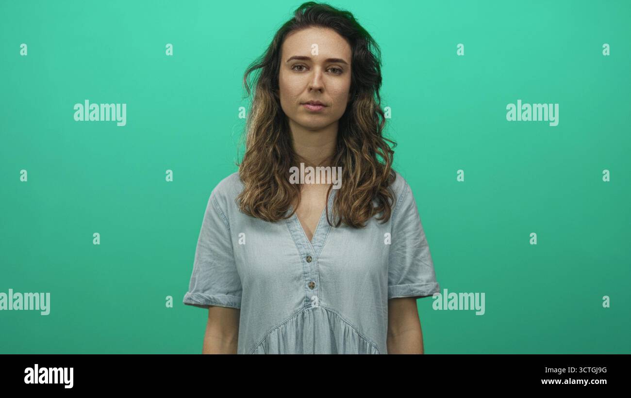 Donna ispanica con abito in denim azzurro incrocia le braccia con espressione neutra in verde studio; rigetto. Foto Stock