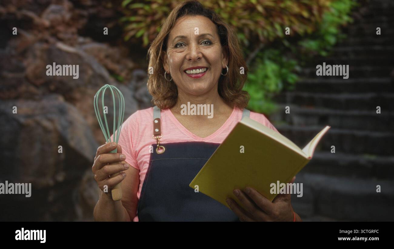 La donna legge il libro di cucina tenendo la frusta su gradini di pietra rocciosa in mezzo al verde della foresta; serenità culinaria attenzione. Foto Stock