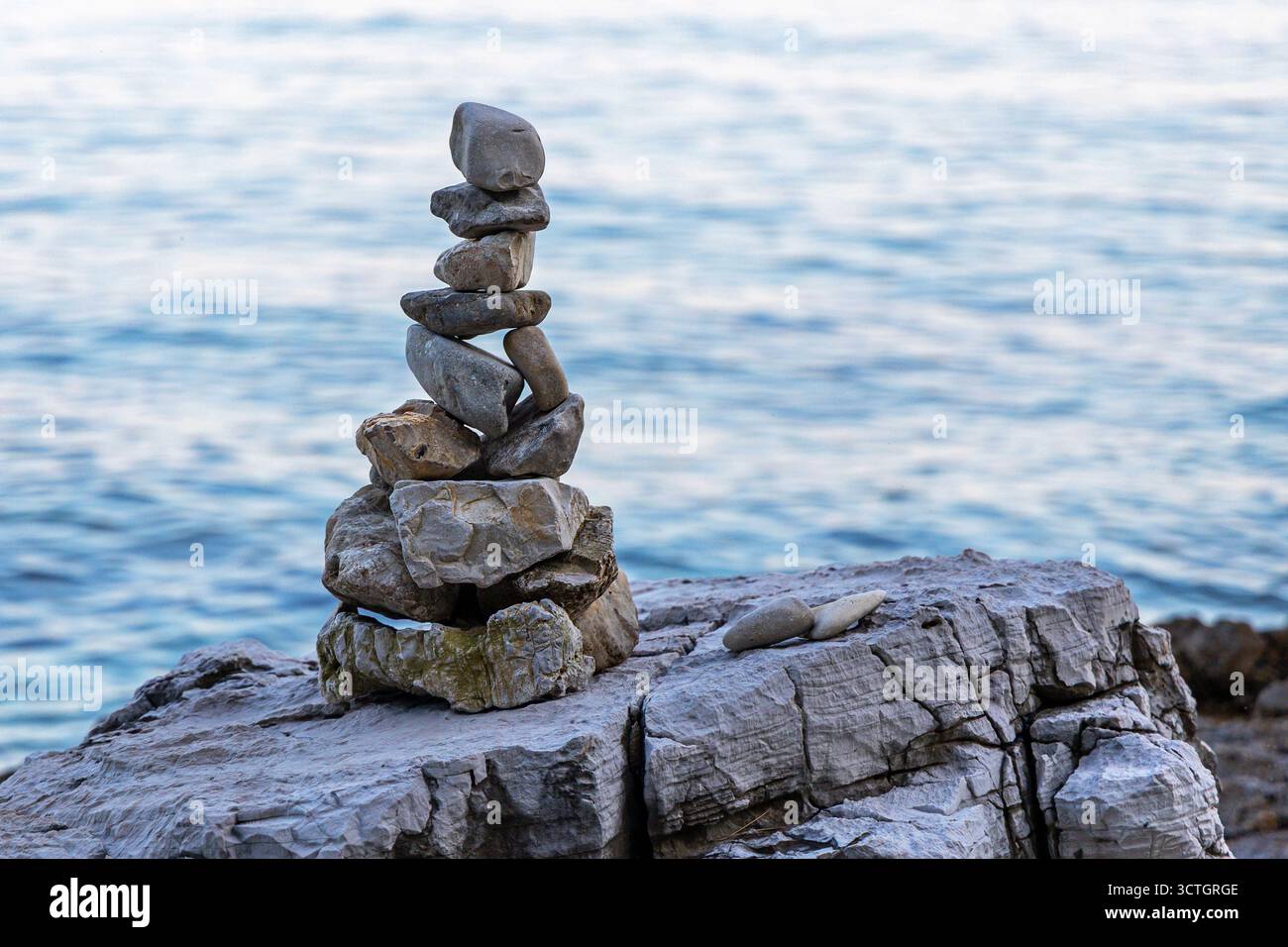 Una pila di pietra bilanciata in riva al mare. Pietre accuratamente impilate formano una torre bilanciata su una riva rocciosa, con calme acque di mare blu sullo sfondo. Foto Stock