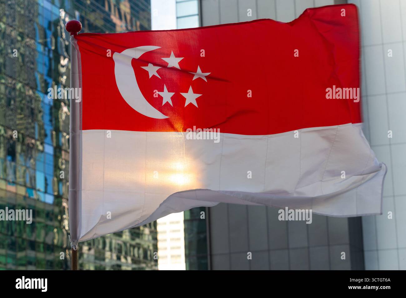 La bandiera nazionale di Singapore ondeggiante, retroilluminata dalla luce del sole che attraversa lo skyline urbano. Design rosso e bianco, a mezzaluna e a stelle. Foto Stock