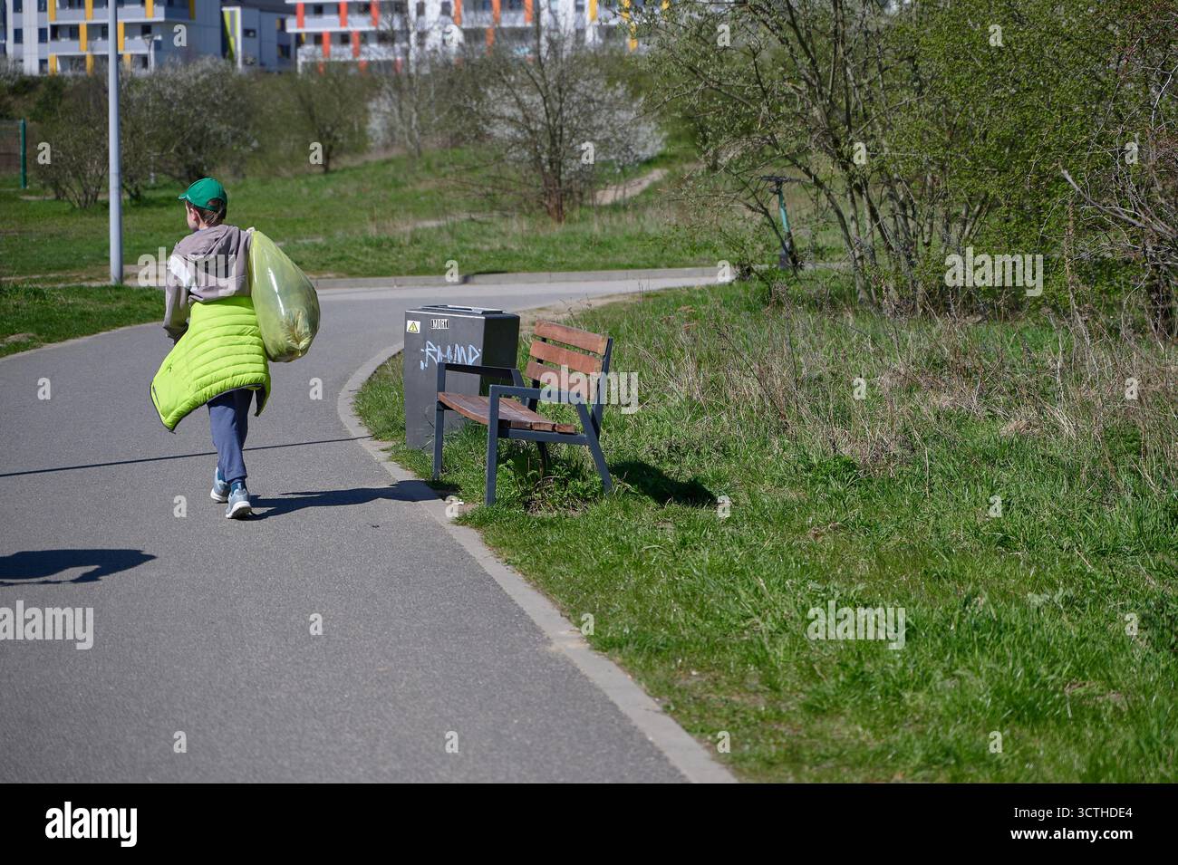 Donna che trasporta una borsa verde per la spazzatura che cammina sul percorso del parco durante la pulizia ambientale, vista posteriore a tutto il corpo alla luce di mezzogiorno Foto Stock