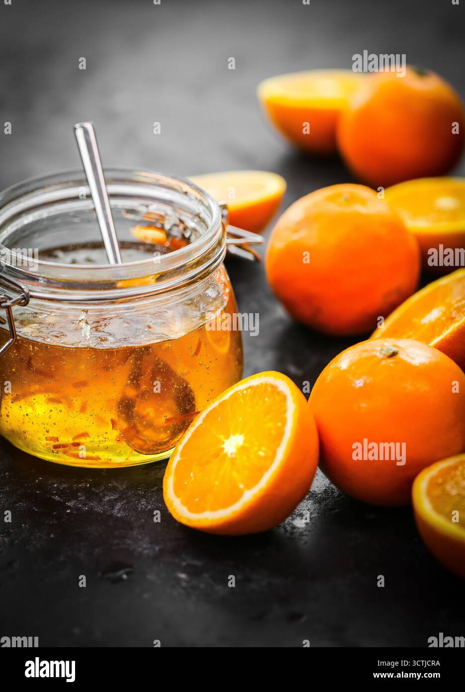 Vaso di vetro con marmellata d'arancia fatta in casa con cucchiaio di metallo e arance tagliate a metà fresche, fotografate in una calda luce naturale per l'uso artigianale di cibo. Foto Stock