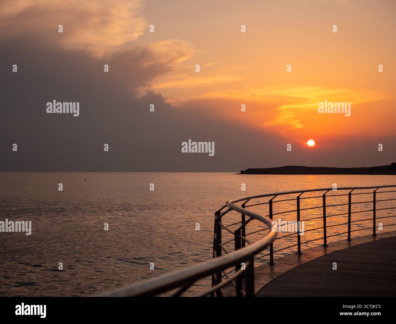 Orbe arancione di un sole che tramonta sul mare Mediterraneo ad Ayia Napa, vista dalla moderna passeggiata con ringhiera in metallo sopra la riva curva e il tramonto Foto Stock