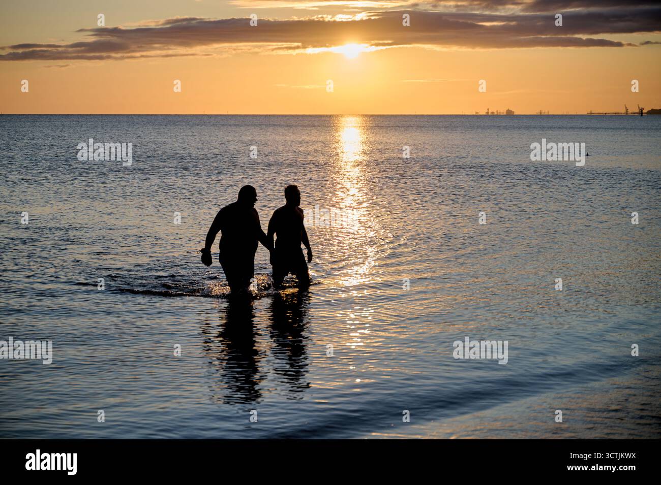 Coppia sagomata che spruzza acqua di mare all'alba, riflesso di luce dorata; orientamento orizzontale, angolo basso, illuminazione naturale per uno stile di vita dinamico Foto Stock