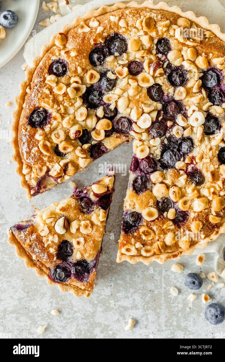 Crostata di frangipane fatta in casa con mirtilli e nocciole con una fetta rimossa, catturata alla luce naturale su una superficie rustica per i temi della panificazione e dei dessert. Foto Stock