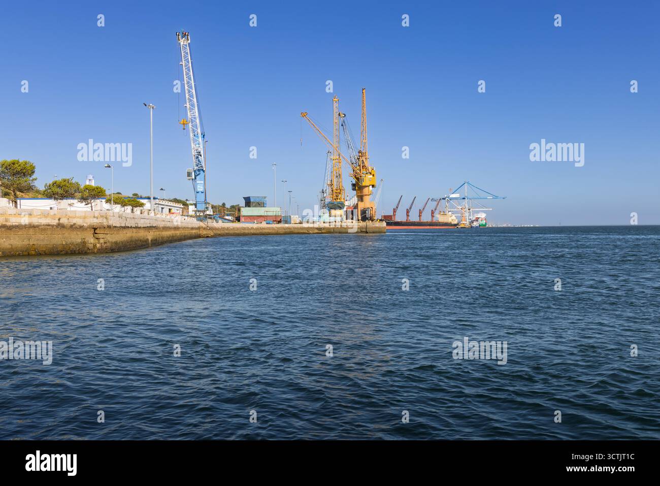 Un'ampia vista del Porto de Setúbal con acque calme in primo piano, che mostra gru cargo e attività industriale sul molo lontano. Setubal, Foto Stock